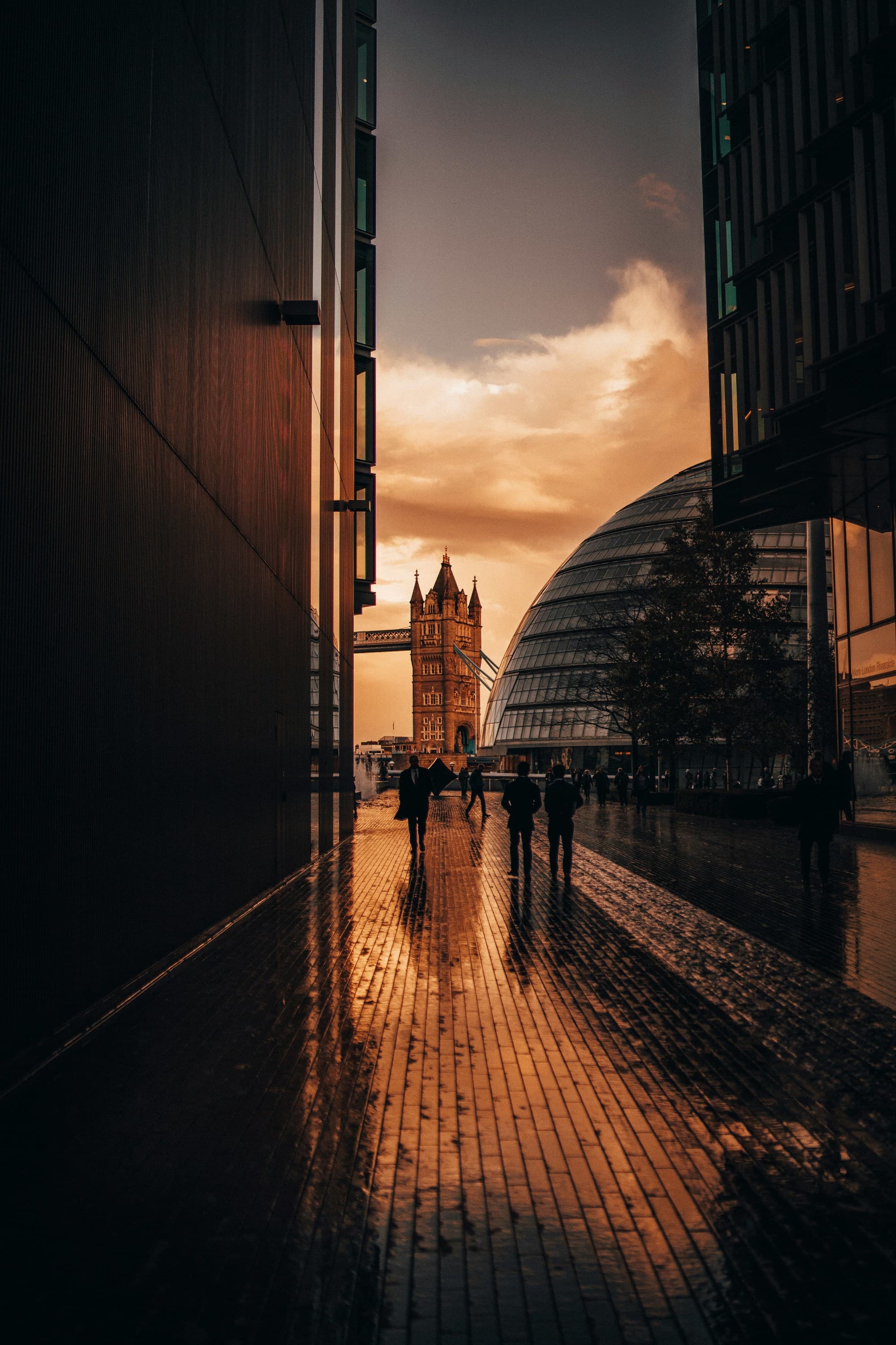A captivating sunset view of the Tower Bridge taken during a London itinerary, enhanced by the reflective sheen of wet pavement and silhouetted figures.