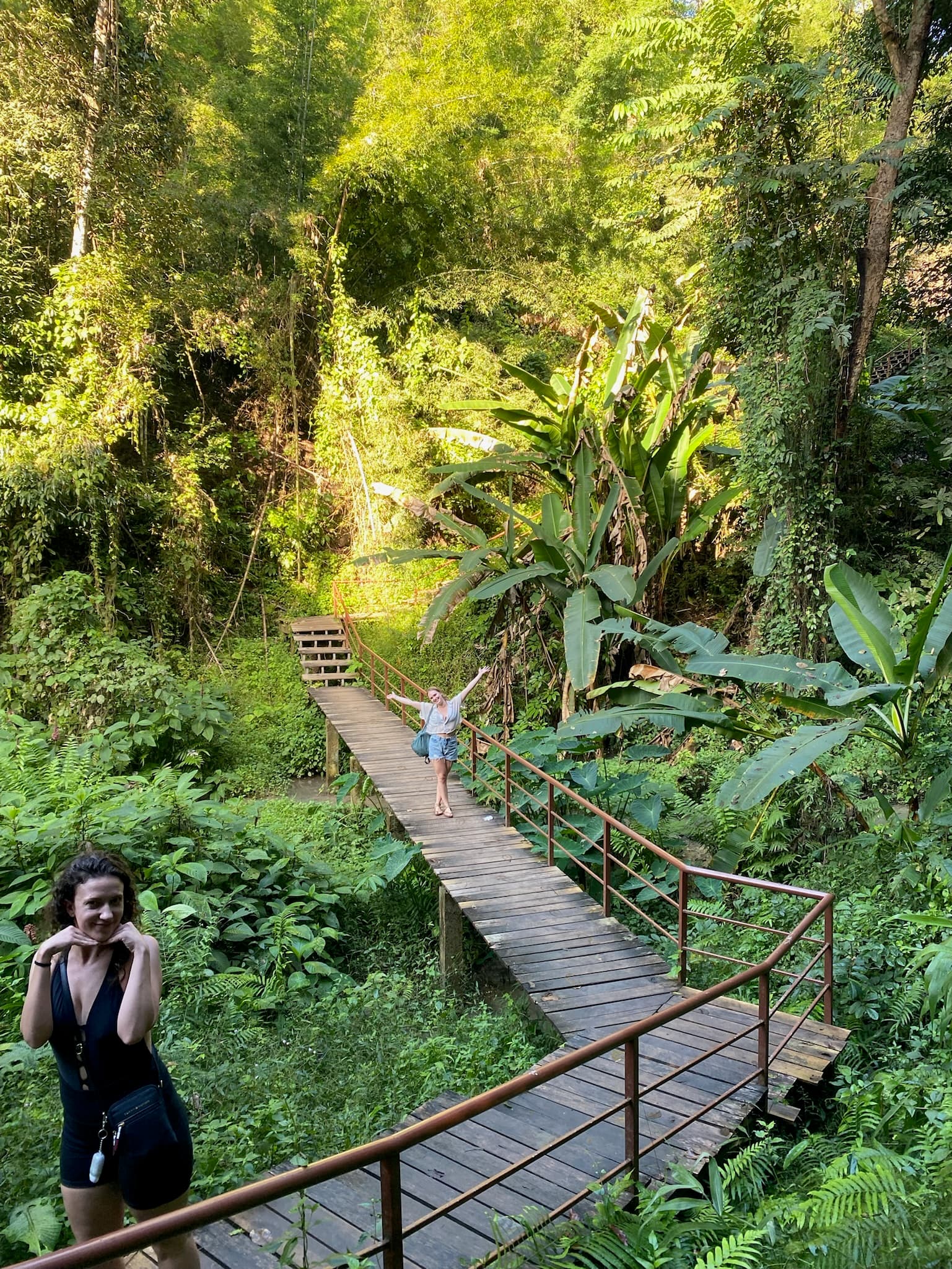 Two women posing on a path surrounded by lush and green plants and trees.
