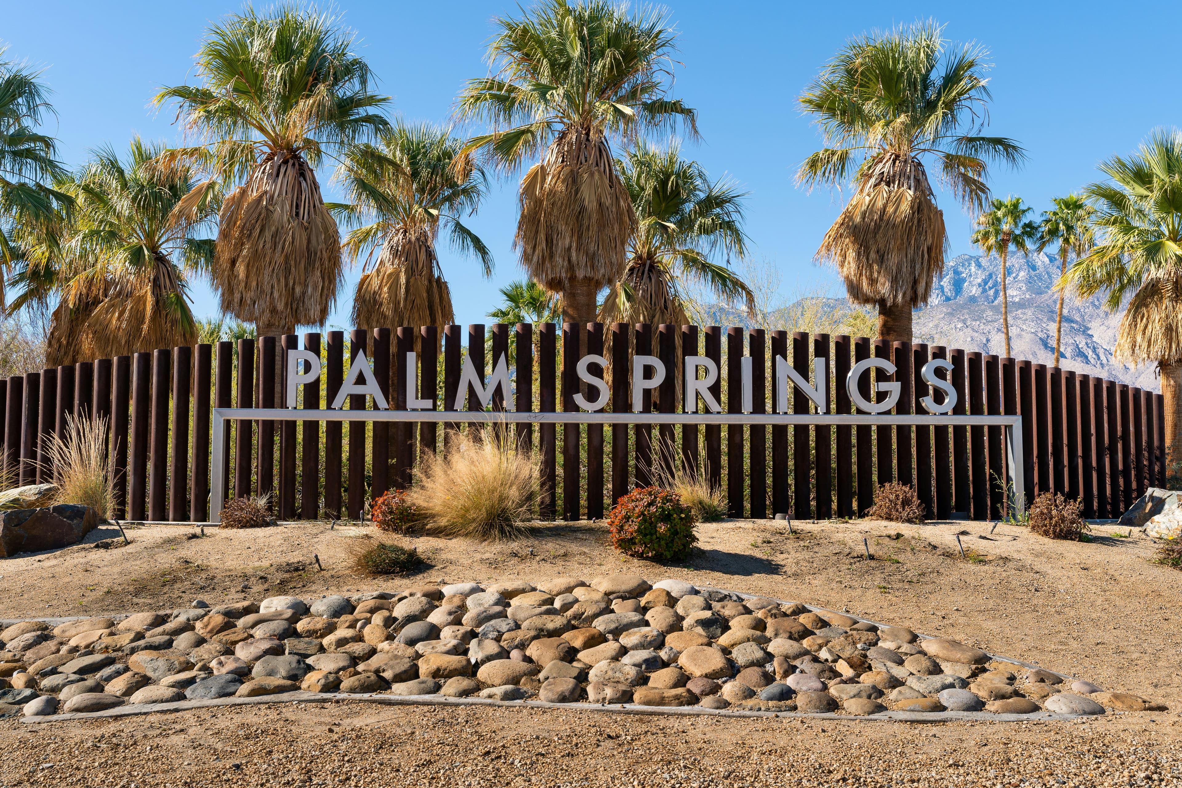 A sign saying "Palm Springs" in big, white lettering, with a fence and palm trees in the background, on a sunny day.