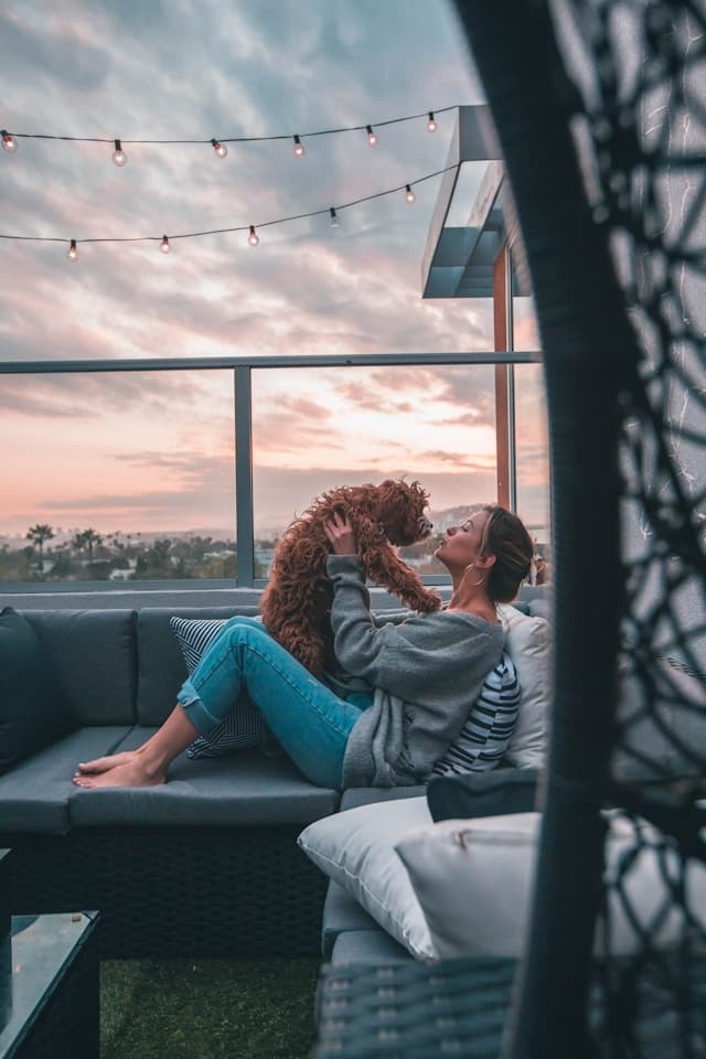 A woman sitting on a balcony on a gray sofa kissing a dog she is holding in her arms. It is sunset and there are string lights above.