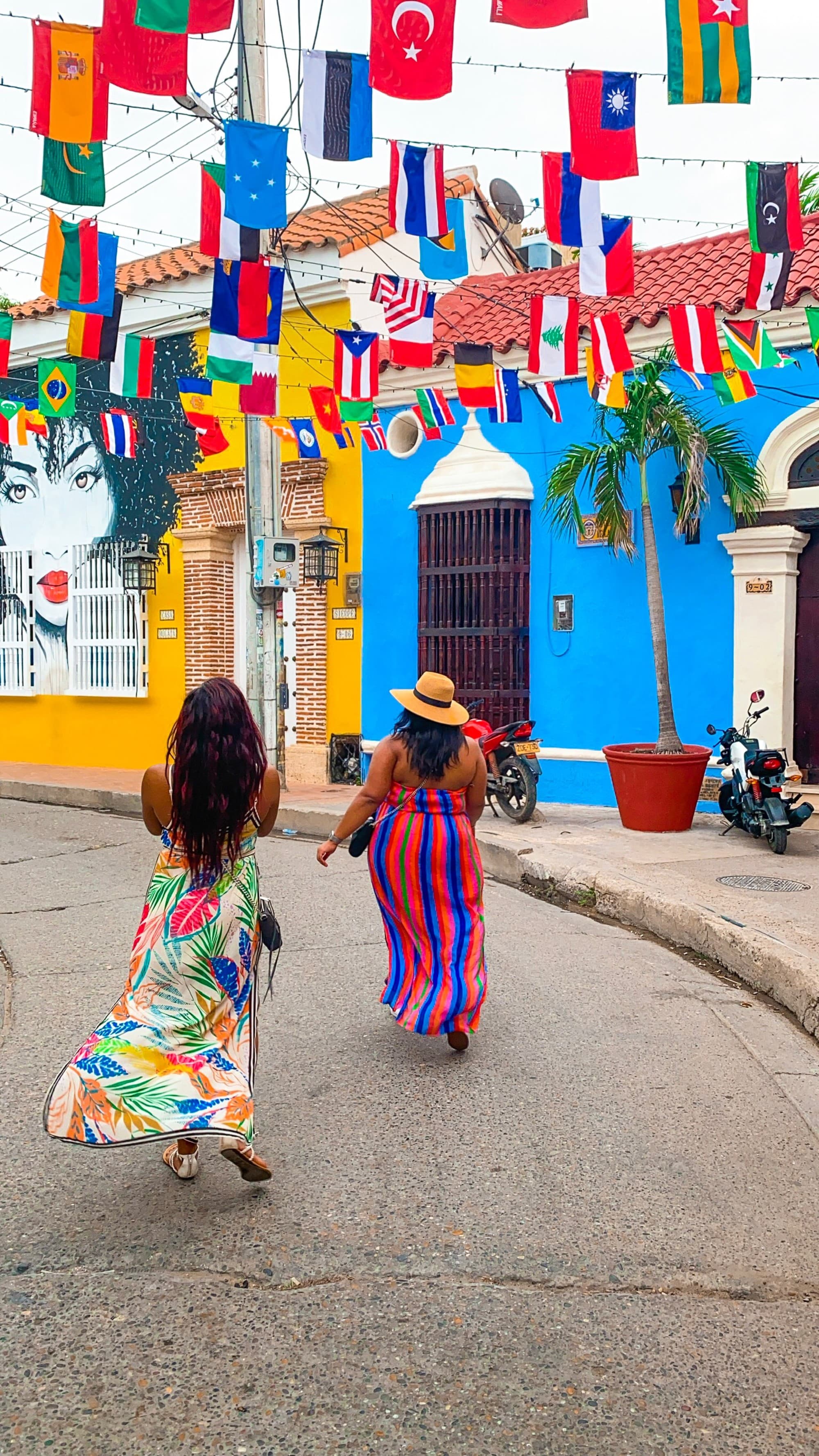 A picture of two people wearing colorful dresses walking down a street with colorful houses and various flags hanging from above at daytime.
