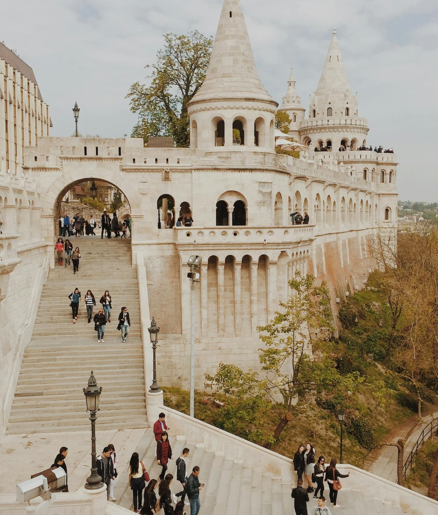 A picture of an old stone building with detailed architecture, steps, towers and trees. There are people walking down the steps and in front of the building.