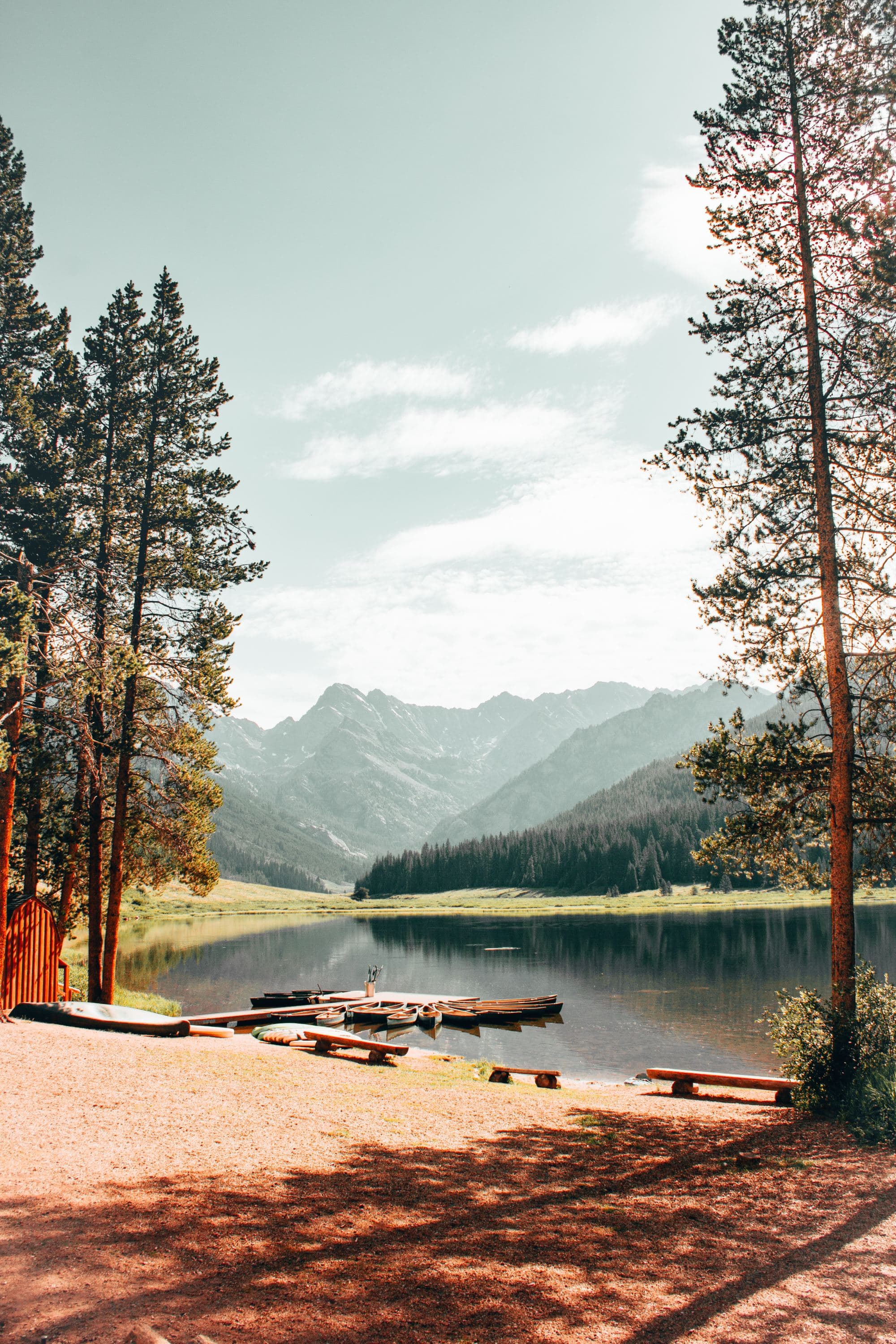 a calm lake with docked canoes at the base of a woodsy mountain region with tall trees framing the scene