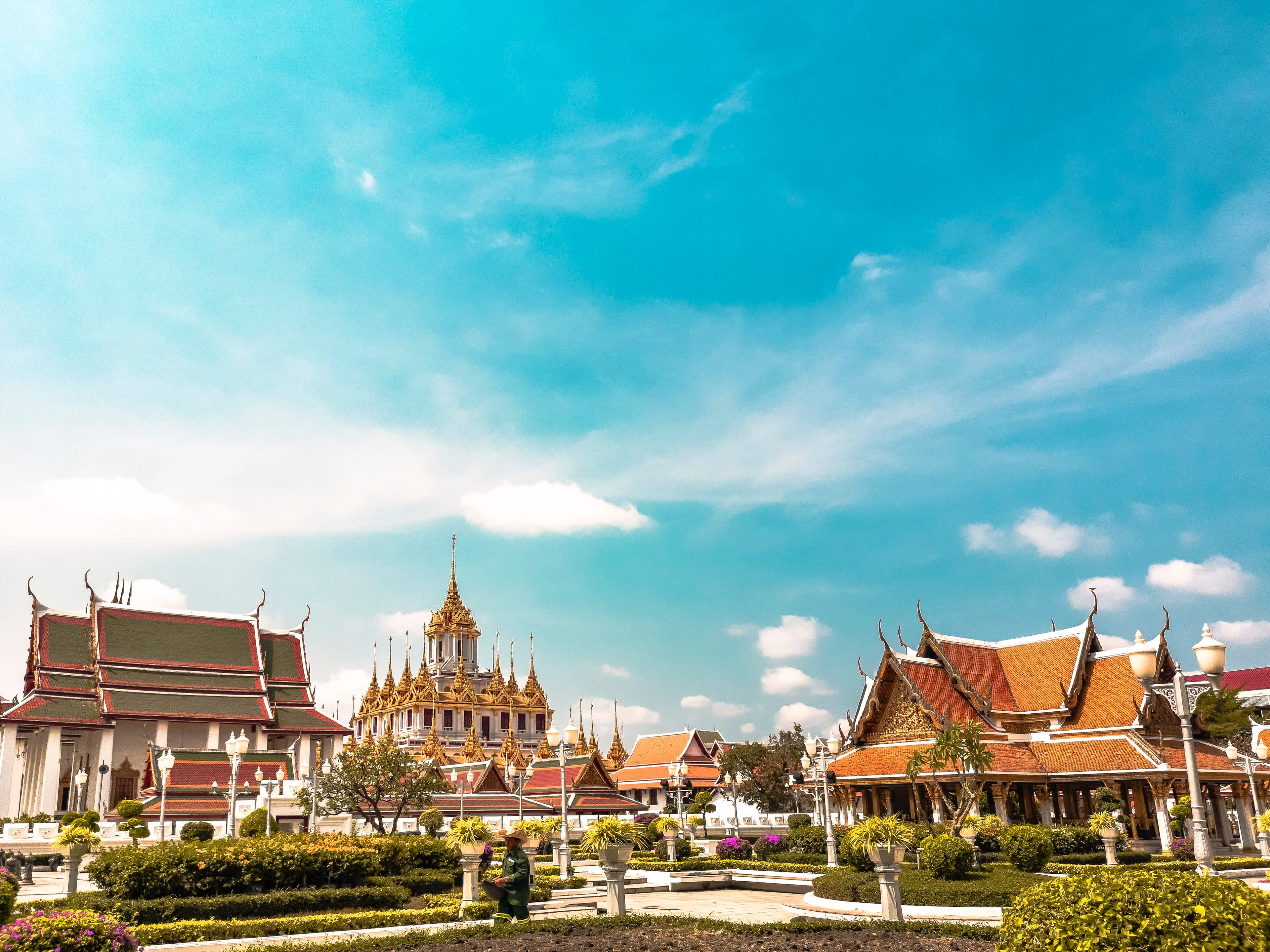 Temples in Bangkok, Thailand with a blue cloudy sky, green groomed bushes, and white paths with people walking on them.