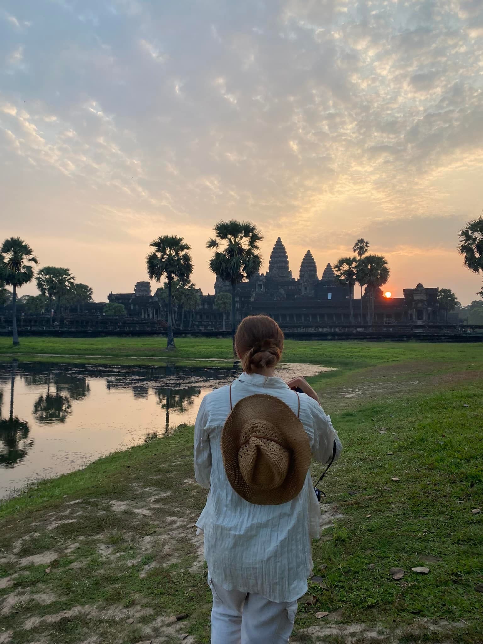 A woman with brown hat facing a building, water body and sunset.