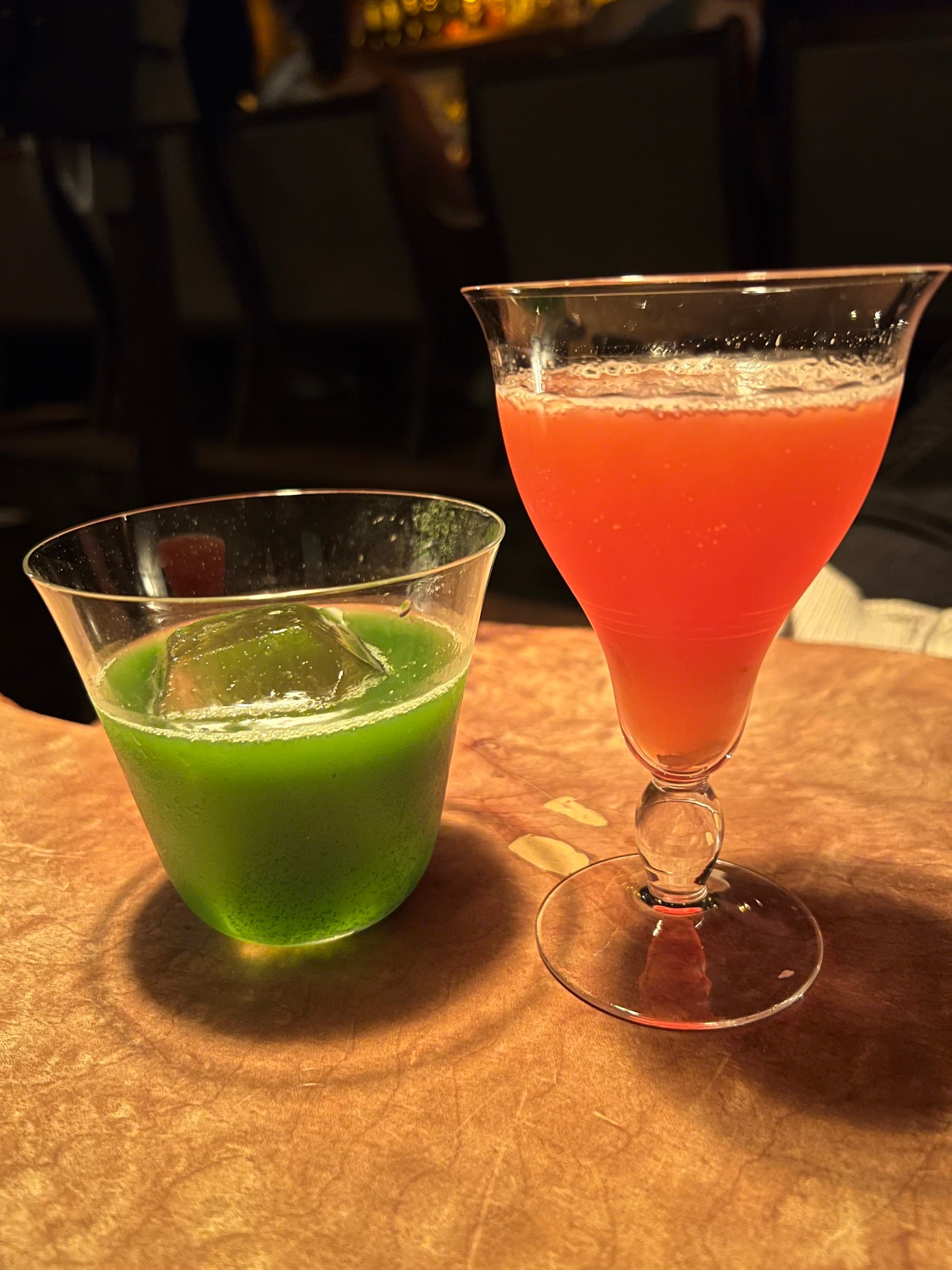 One orange and one green cocktail sitting on a bar.