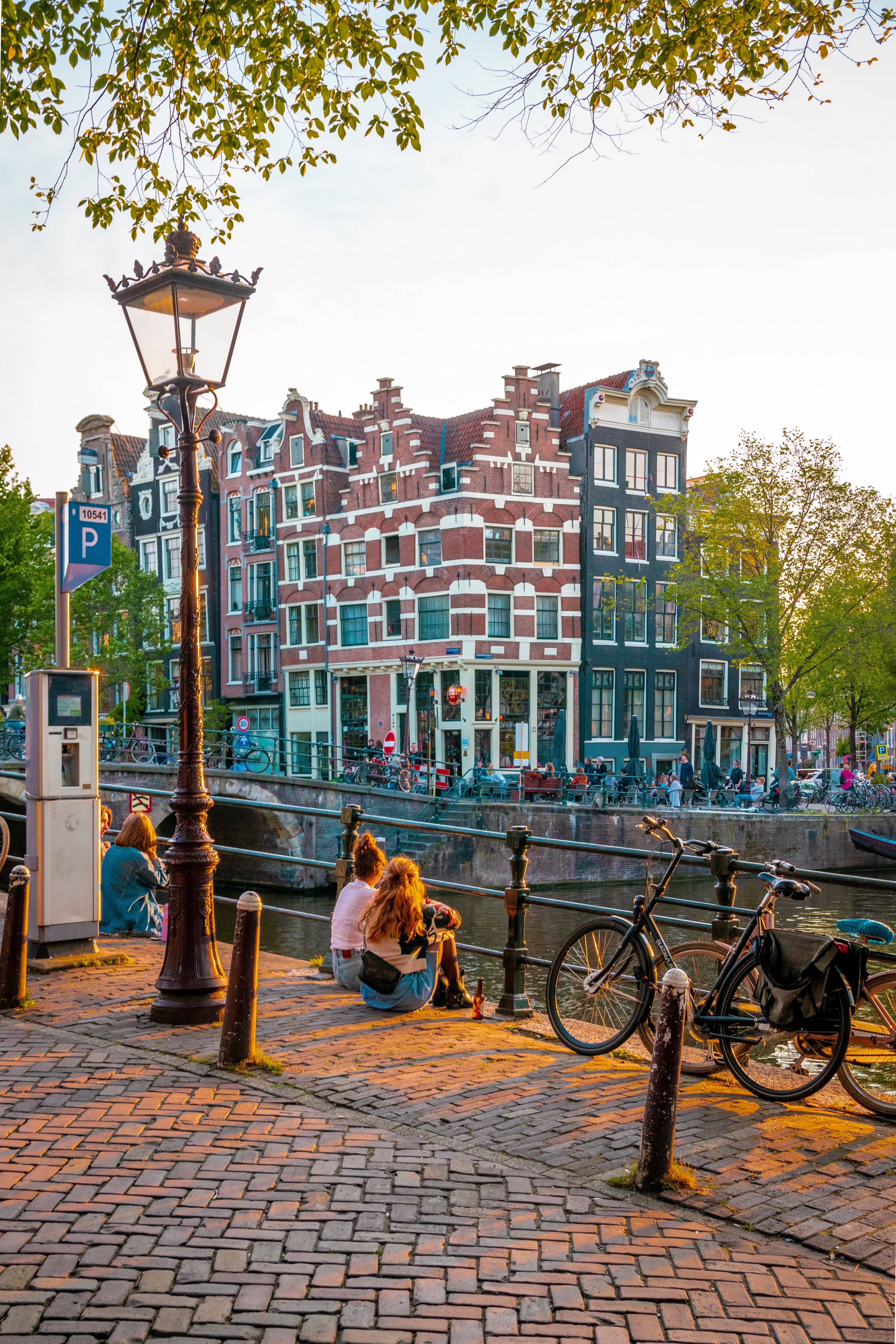 People sitting next to bikes and canal with buildings in the background during daytime