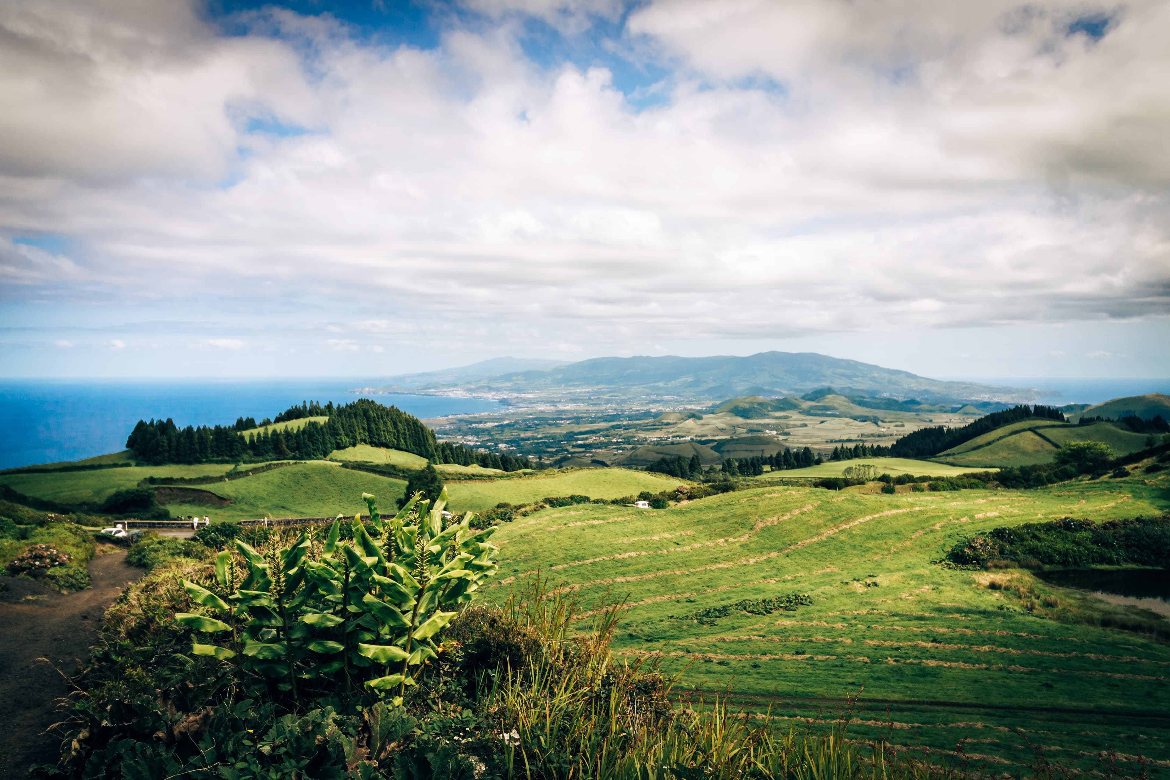 green rolling hills in Azores Portugal with tall trees and grassy bushes and blue ocean water with a blue cloudy sky