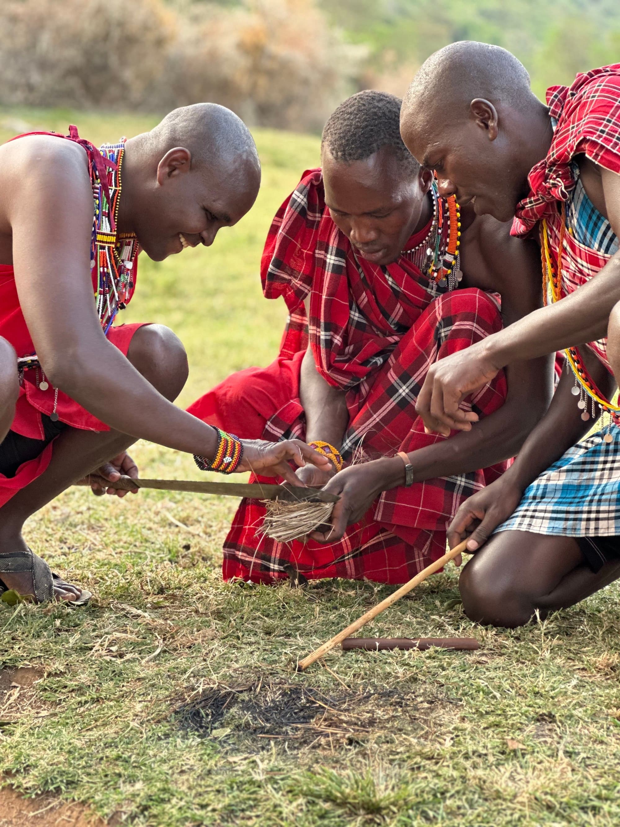 a group of Maasai people glad in red plaid garments crouch down to build a fire