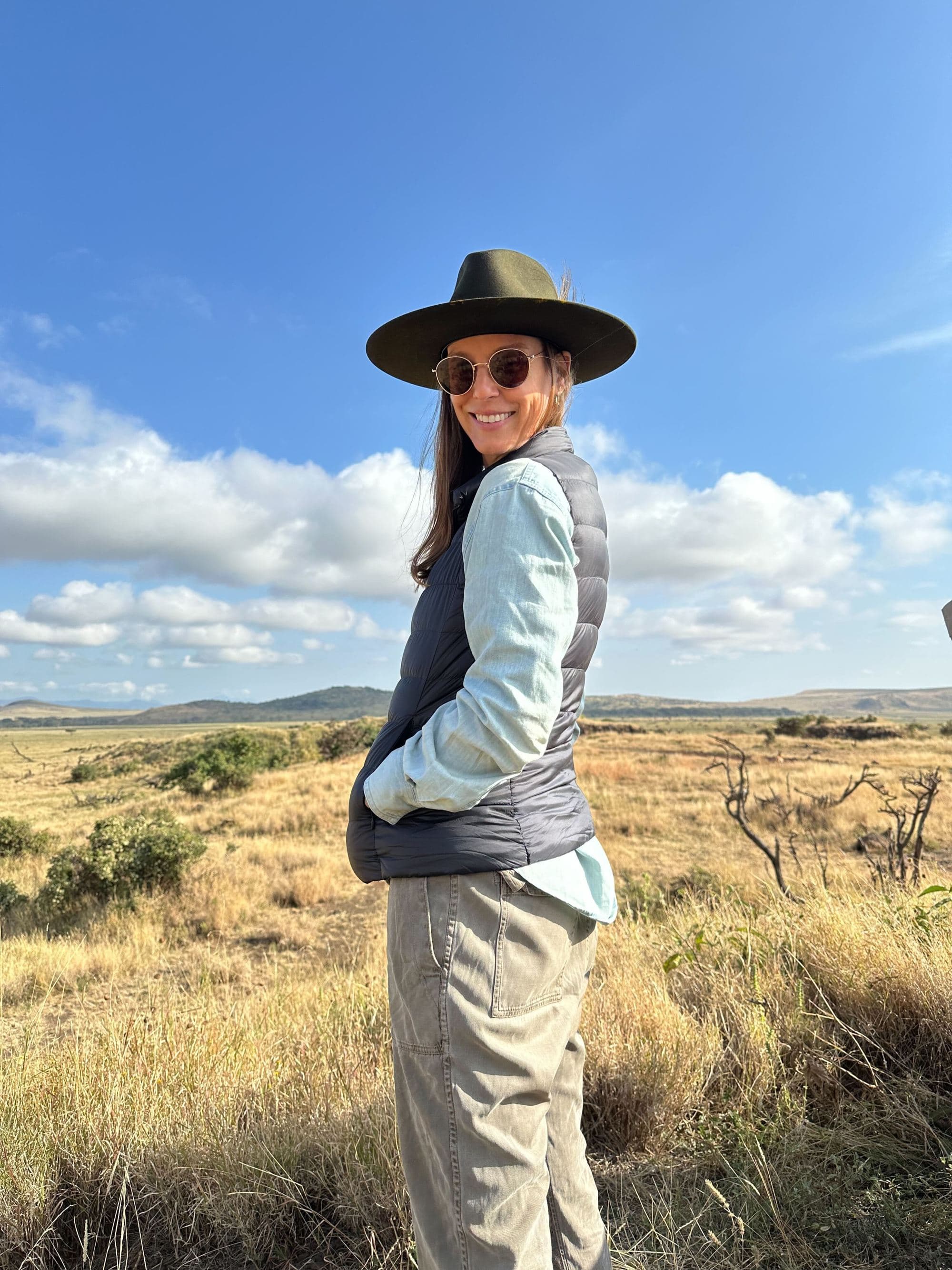 a woman in a black hat and sunglasses stands on a grassy African plain