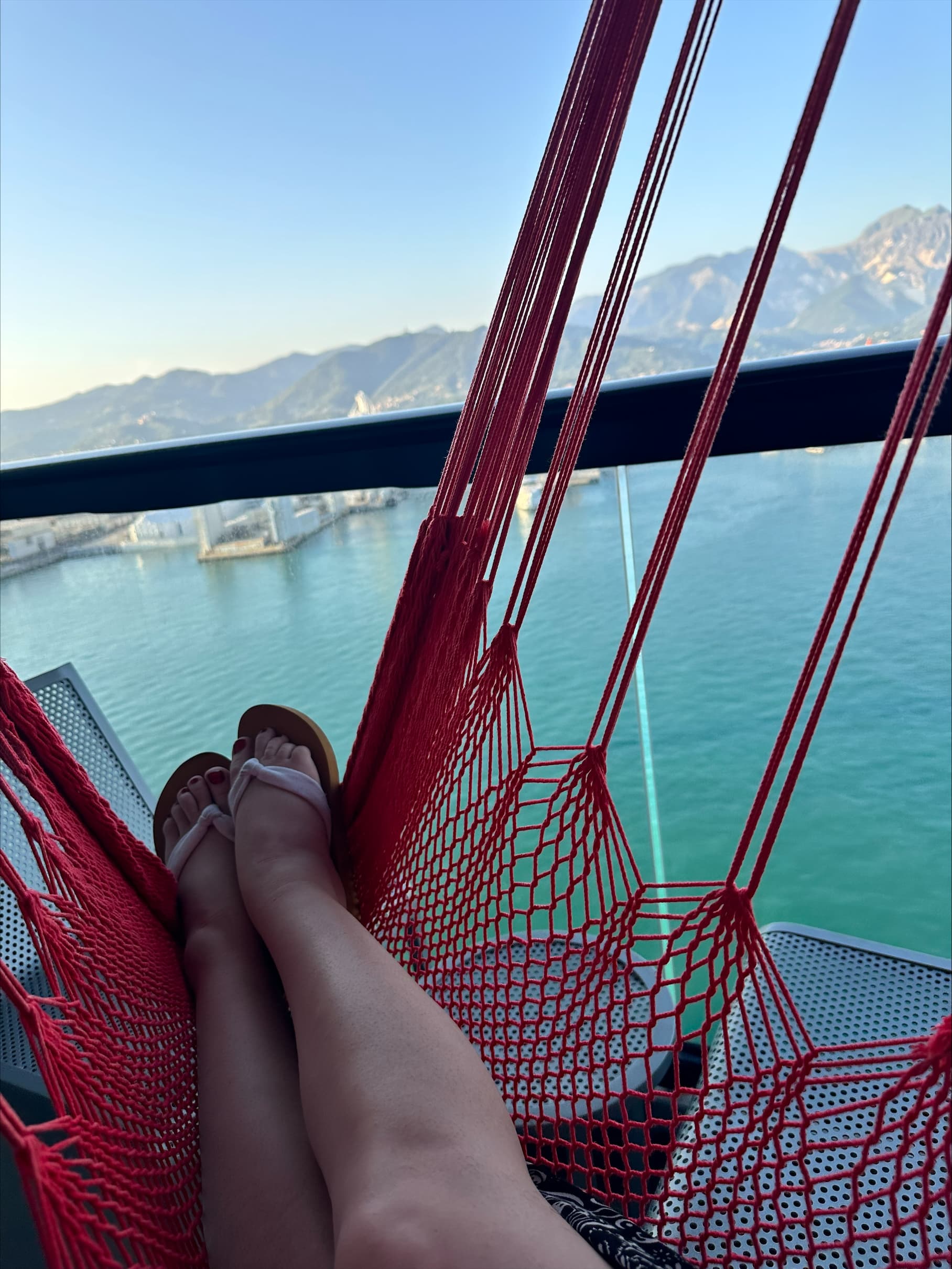 A person's legs resting in a red swing with a view of the water during the daytime