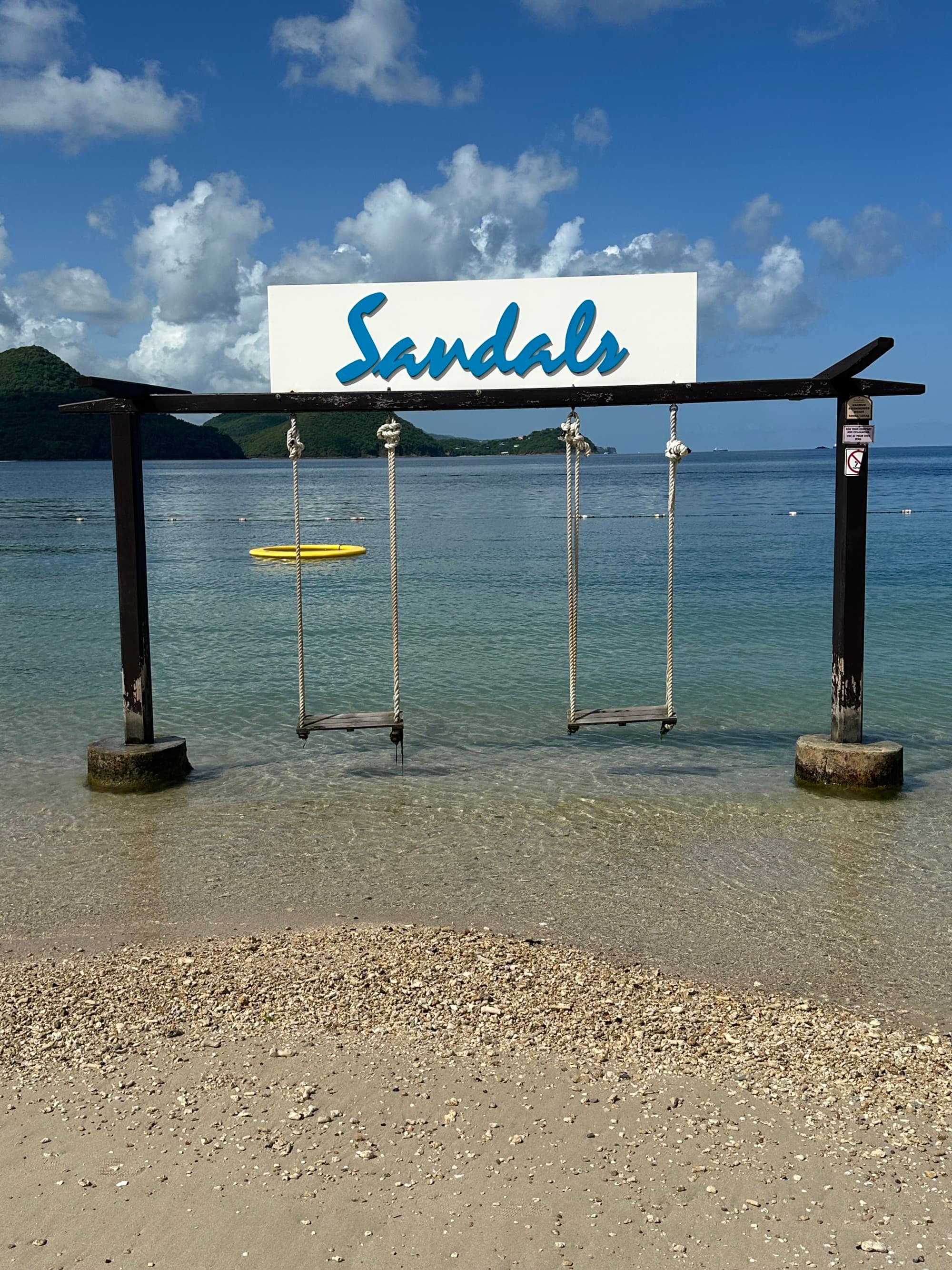A sign reading "Sandals" above a swing set in the water on the beach