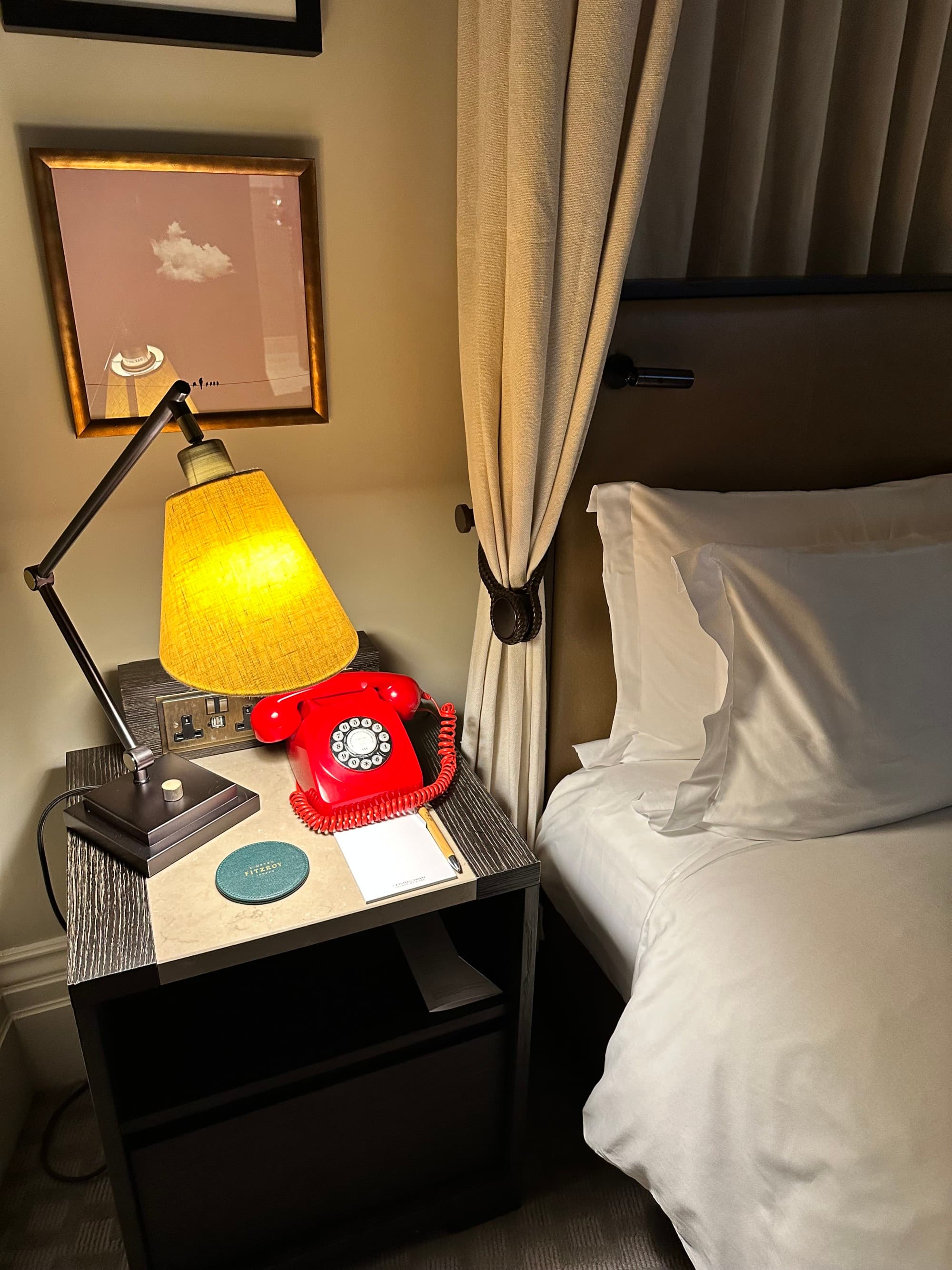 Hotel bed with white linens next to a nightstand with a red rotary phone and a yellow table lamp.