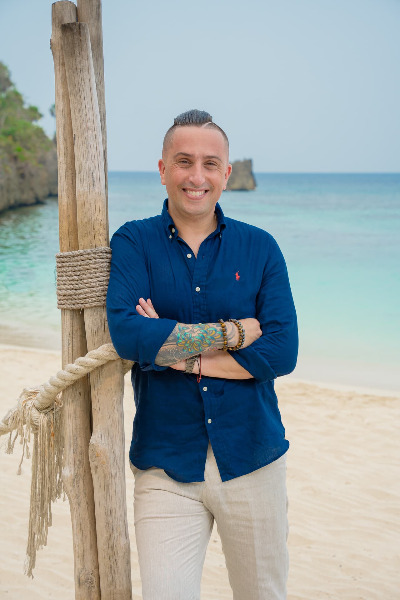 man in blue shirt leading on a wooden post with beach and sea in the background