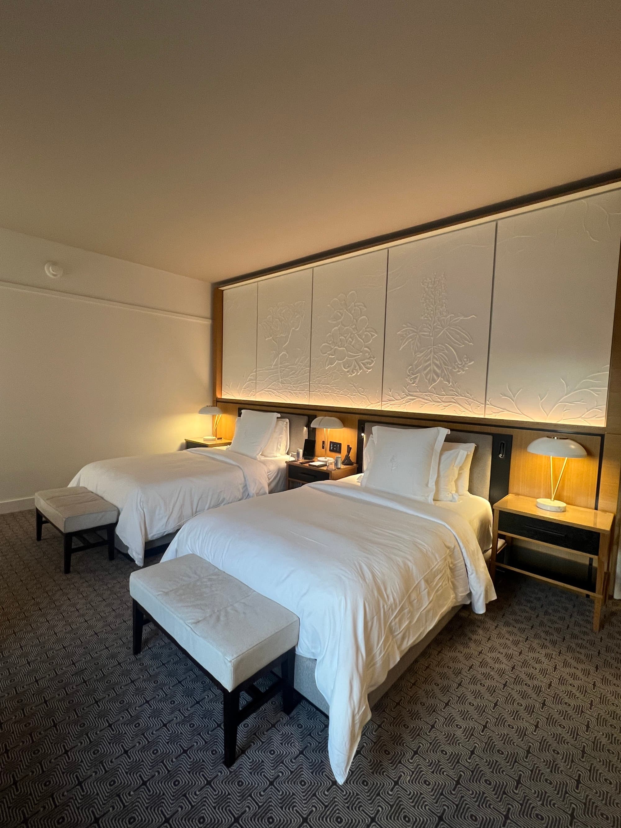 A view of a serene hotel room with white walls, bedding and benches at the end of each bed.