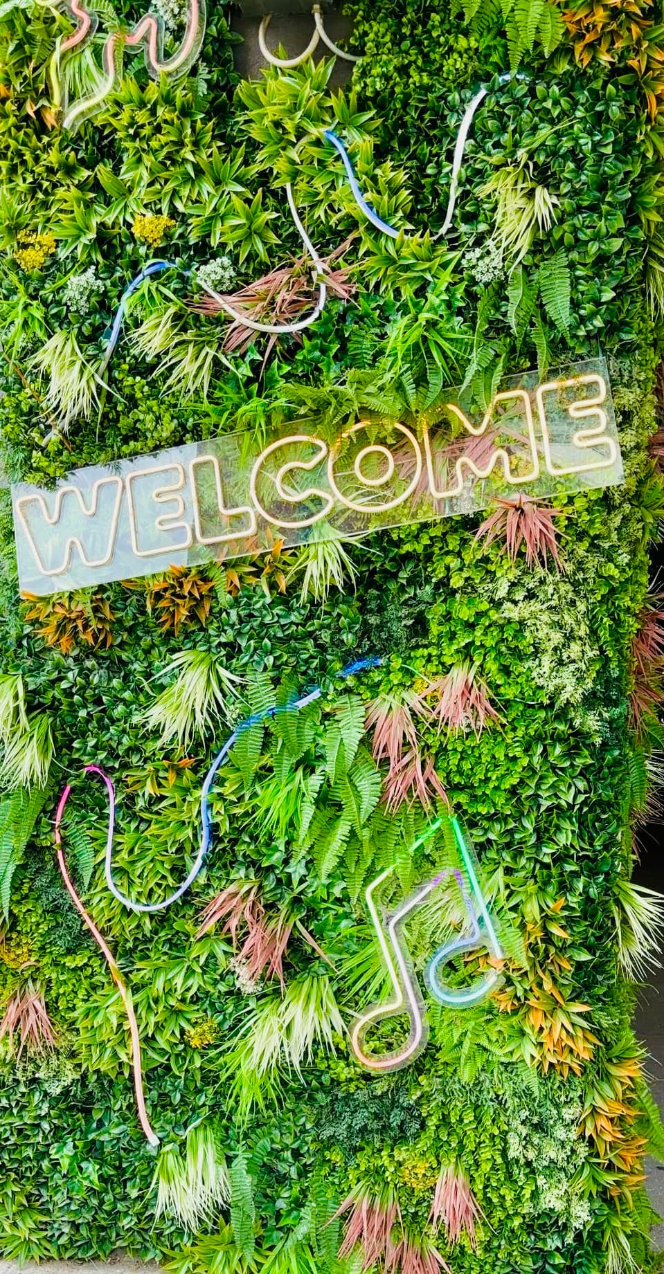 Outside wall with plants covering it and a neon sign that says "Welcome."