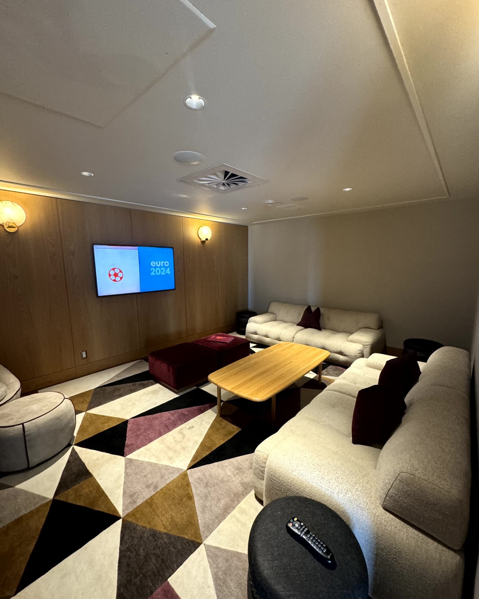 A view of a common space with cream couches, a wooden table and TV on the wall. The carpet is black, grey, brown and white geometric shapes. Accommodations - Fairmont Gold 2 - Rondene Grinam