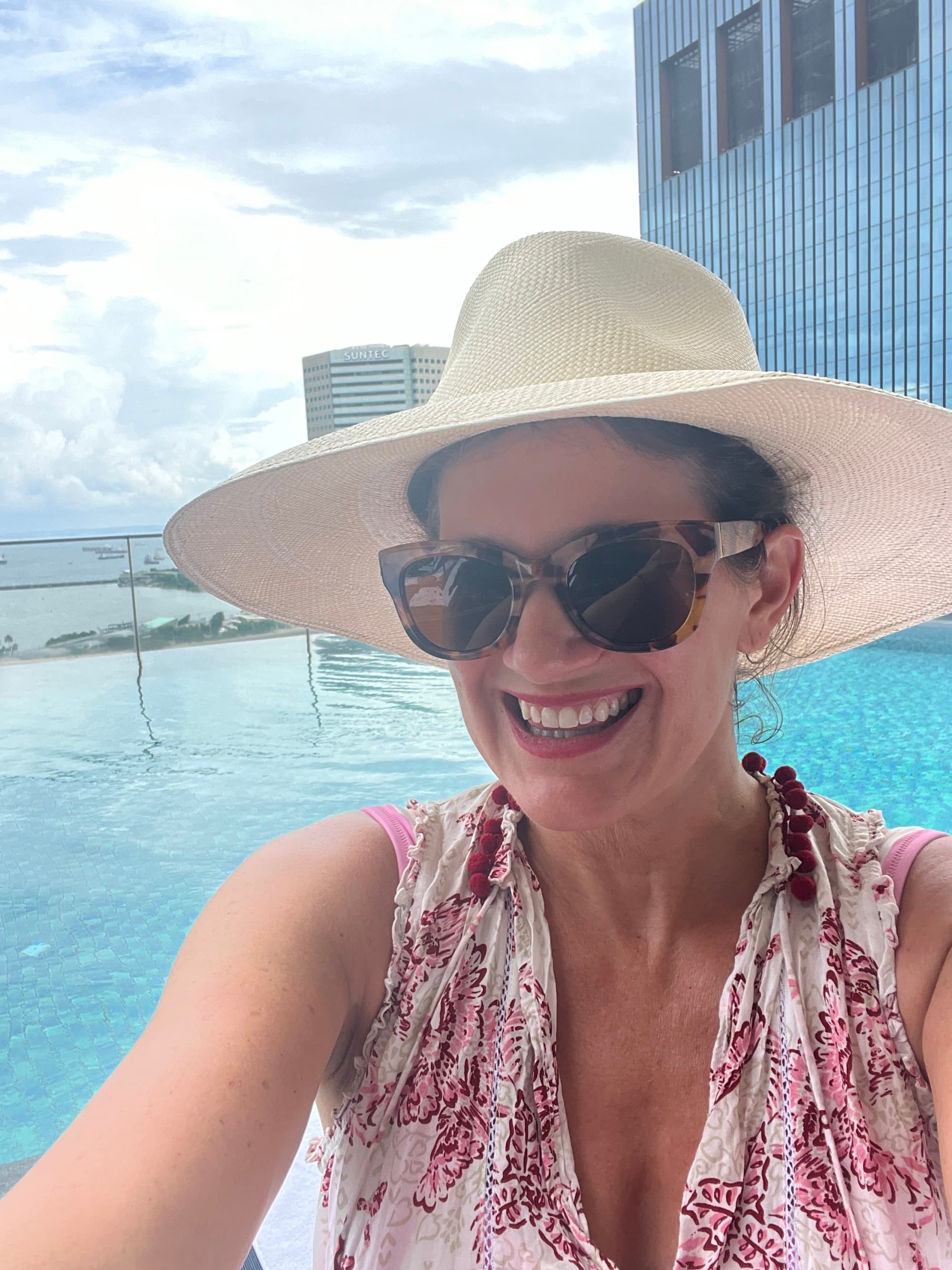 Selfie of a woman wearing sunglasses and a broad-brim hat in a pool.