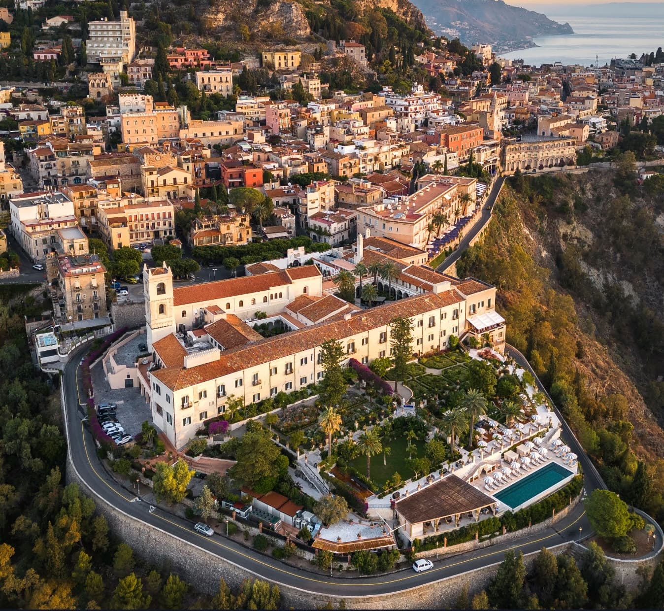Aerial view of an Italian hotel on a cliff with cream stucco exterior and red tile room plus a pool