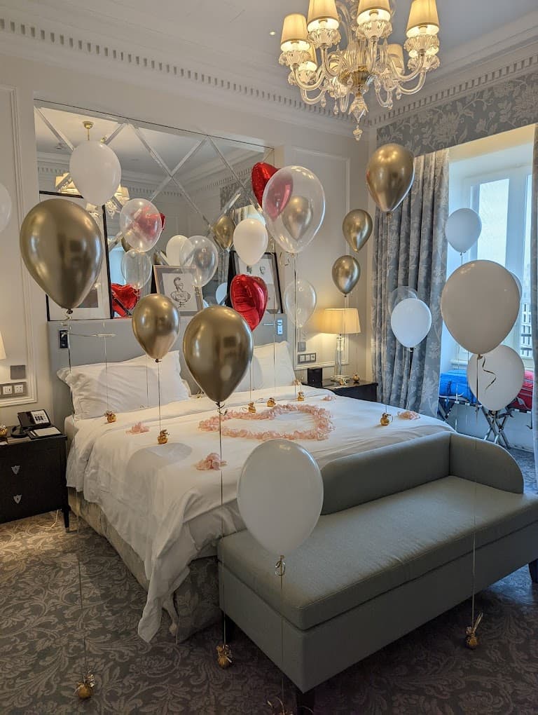 A hotel room with a white bed topped with a heart made of flowers and gold and pink balloons.