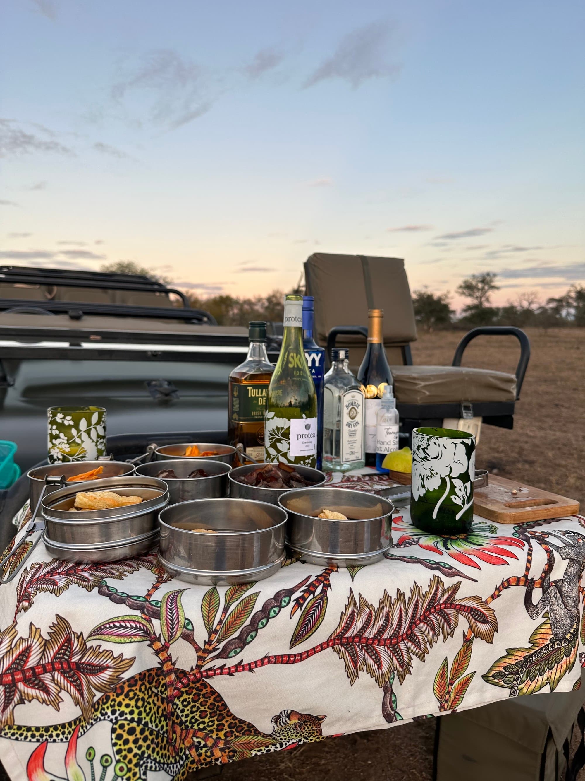 Sundowner Spread, with bowls and bottles of wine on a picnic table covered with a tablecloth.