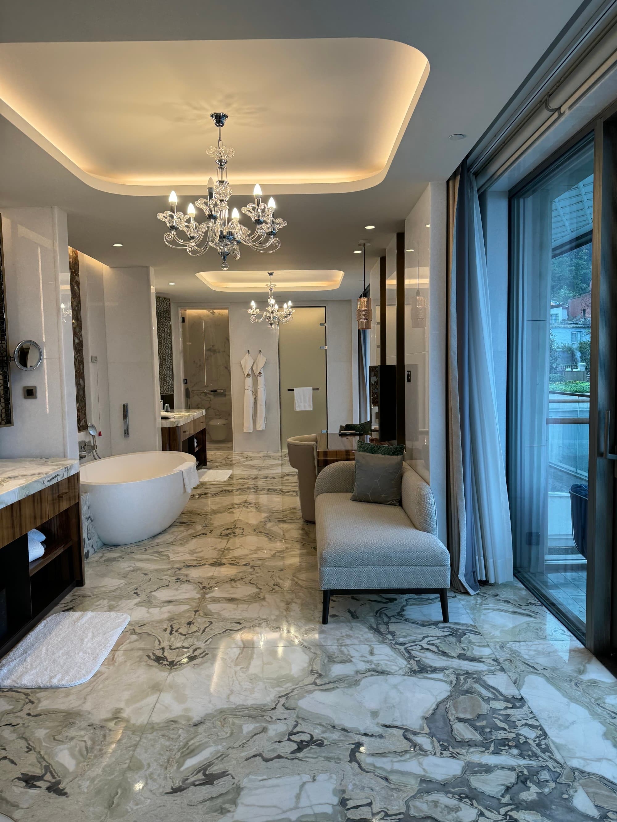 A luxurious and spacious bathroom suite with a marble floor, ceramic tub, a chez lounge, double sinks, and toilet behind it's own door.