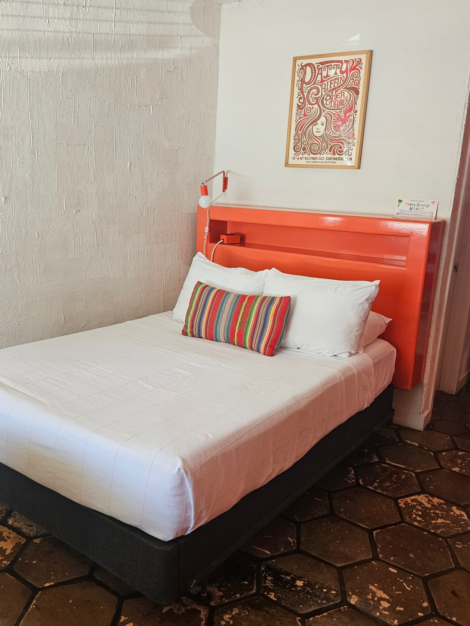 A white bed with a colorful accent pillow and orange headboard on top of a tile floor - Nicole Diez