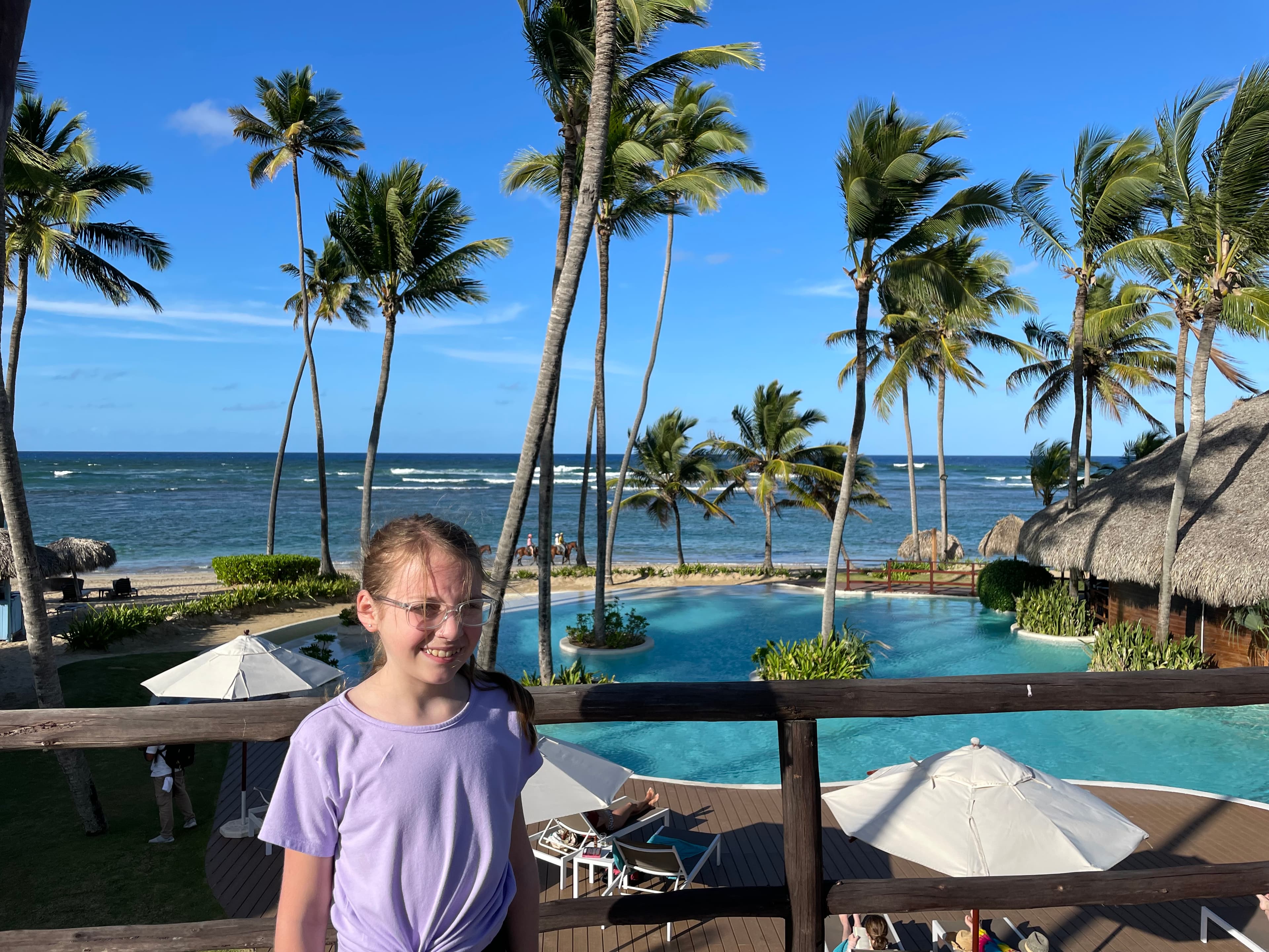 A young girl standing on a balcony overlooking the resort and pool area with palm trees swaying in the wind