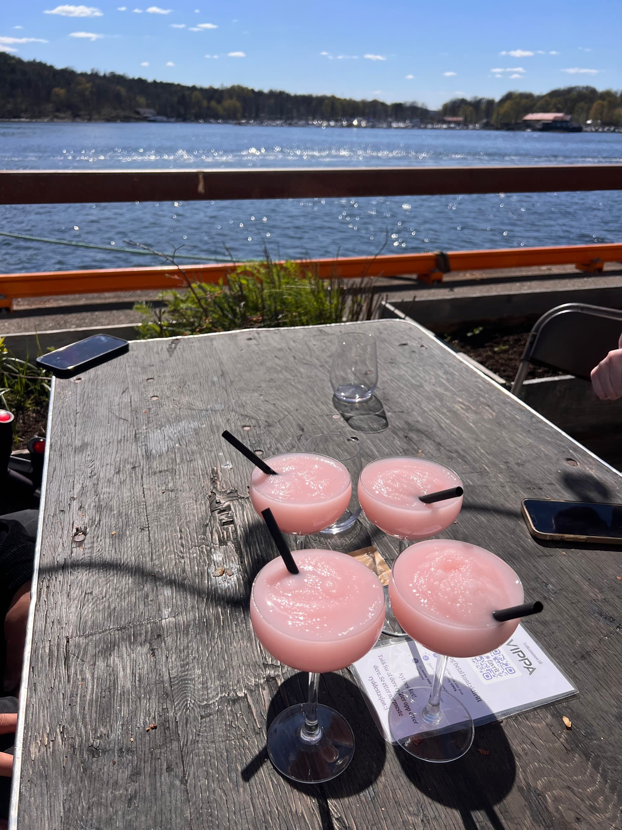 Four pink cocktails on a table in front of a body of water
