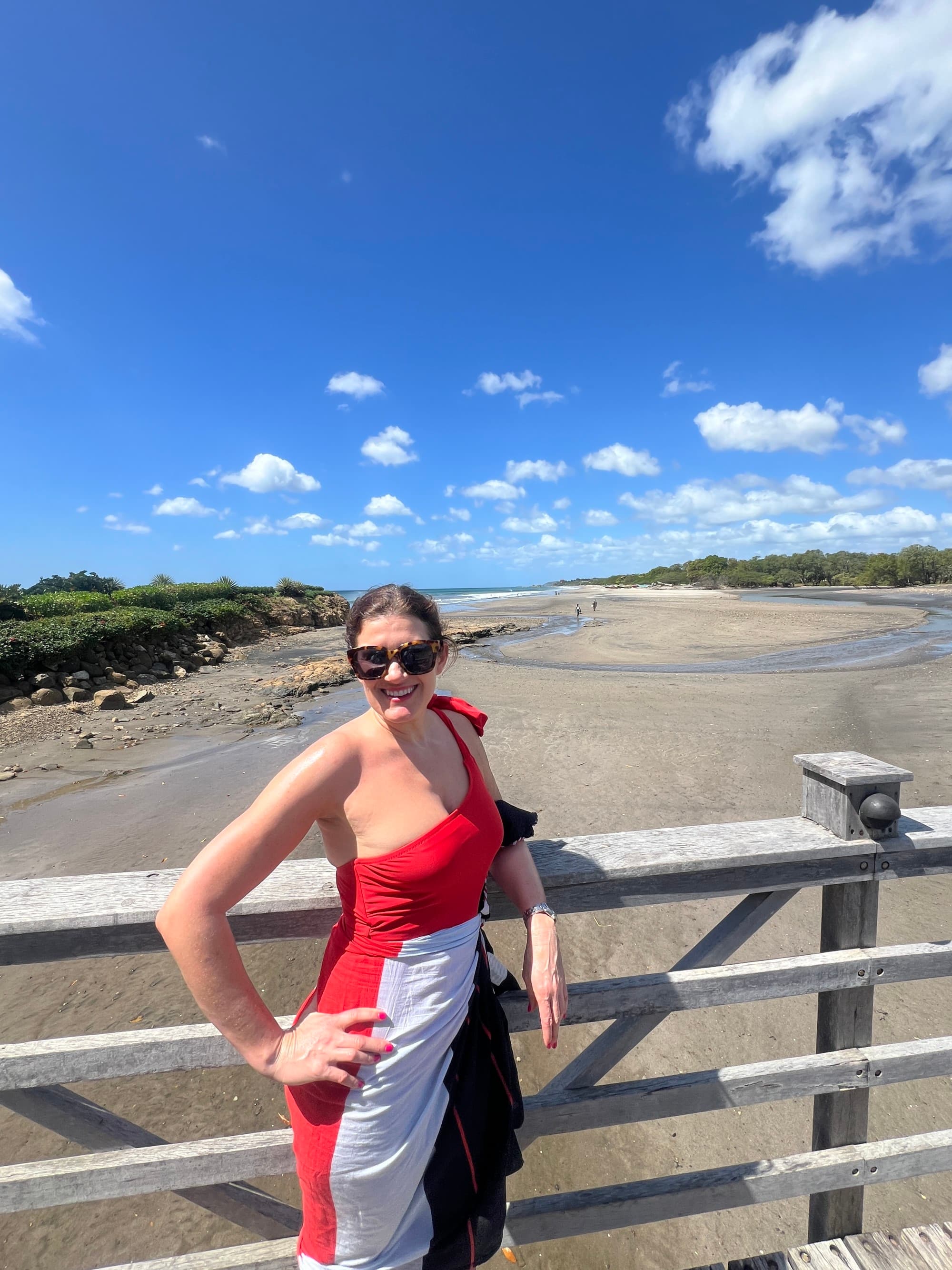 Travel advisor Emily standing on a wooden deck in a red, white and black dress in front of a beautiful beach
