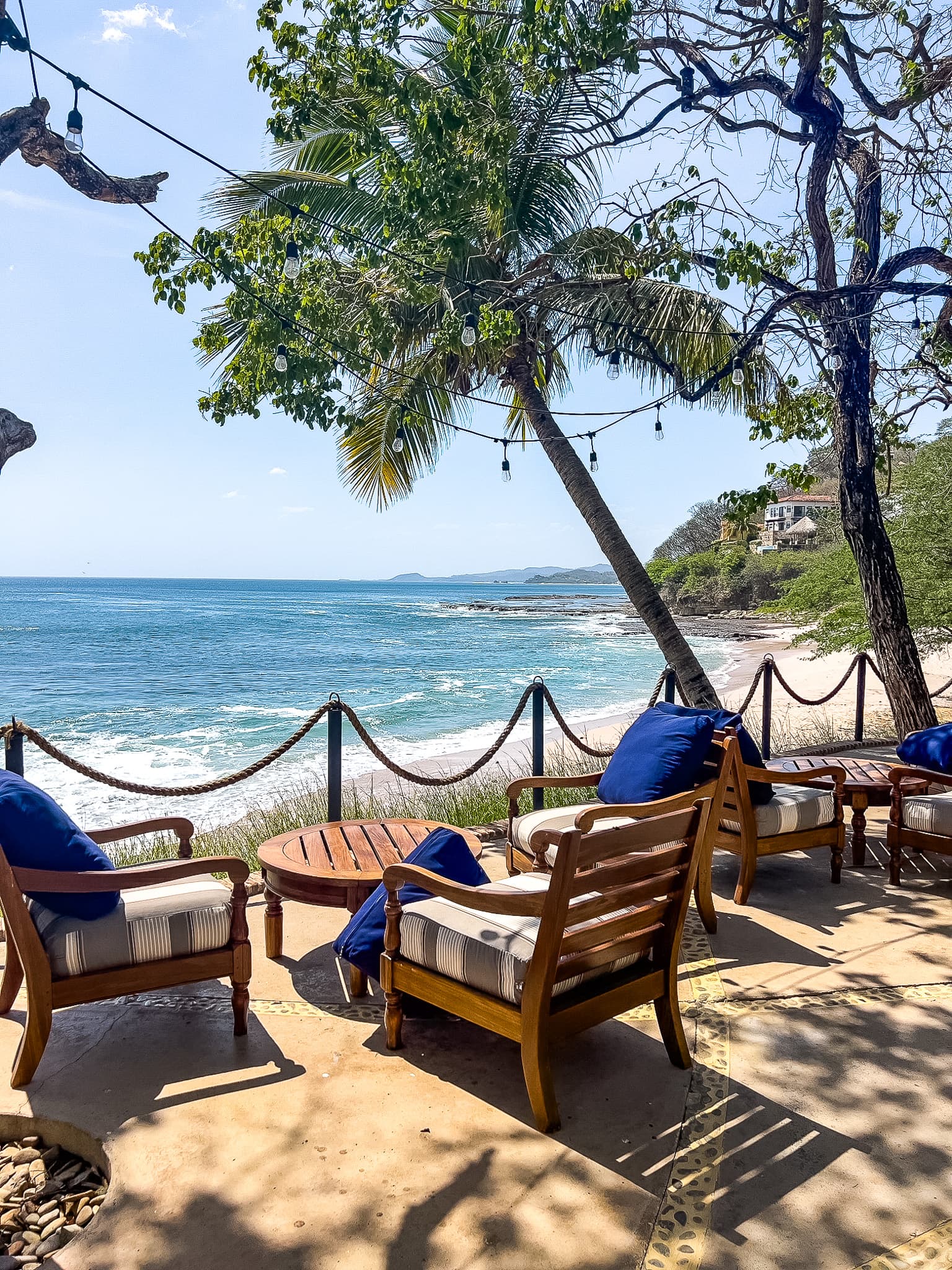 An outdoor lounge area composed of wooden chairs, soft cushions and a wooden table, in front of the beach.