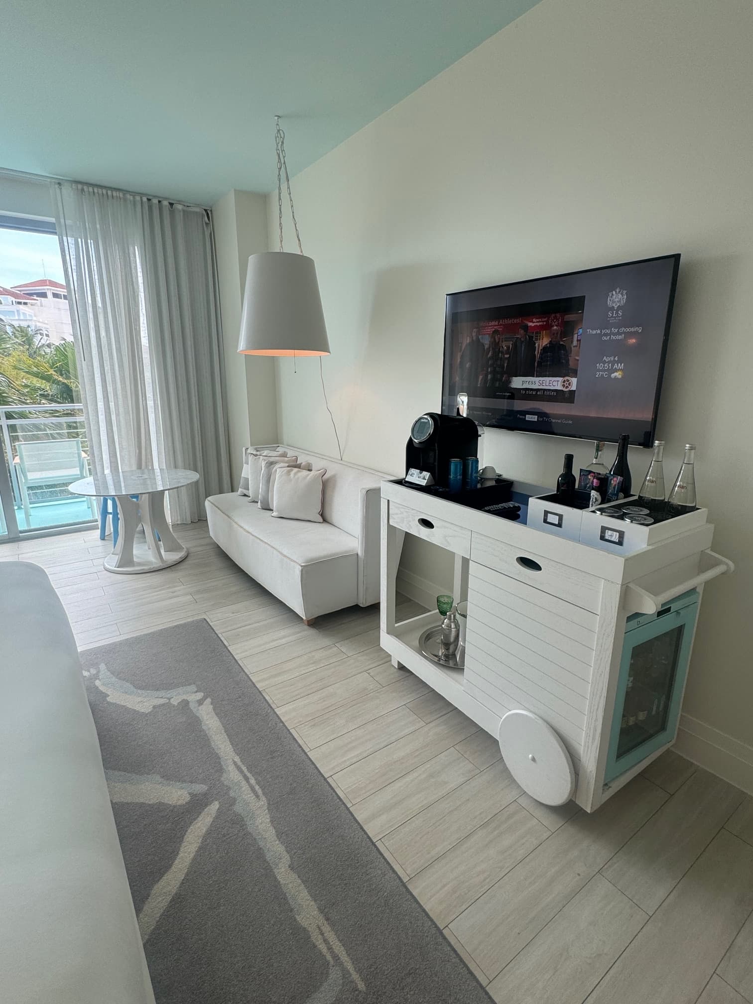 An image of a hotel suite with lounge seating, hanging light, side table, bar cart, television mounted onto a wall and a rug with windows and curtains in the background.