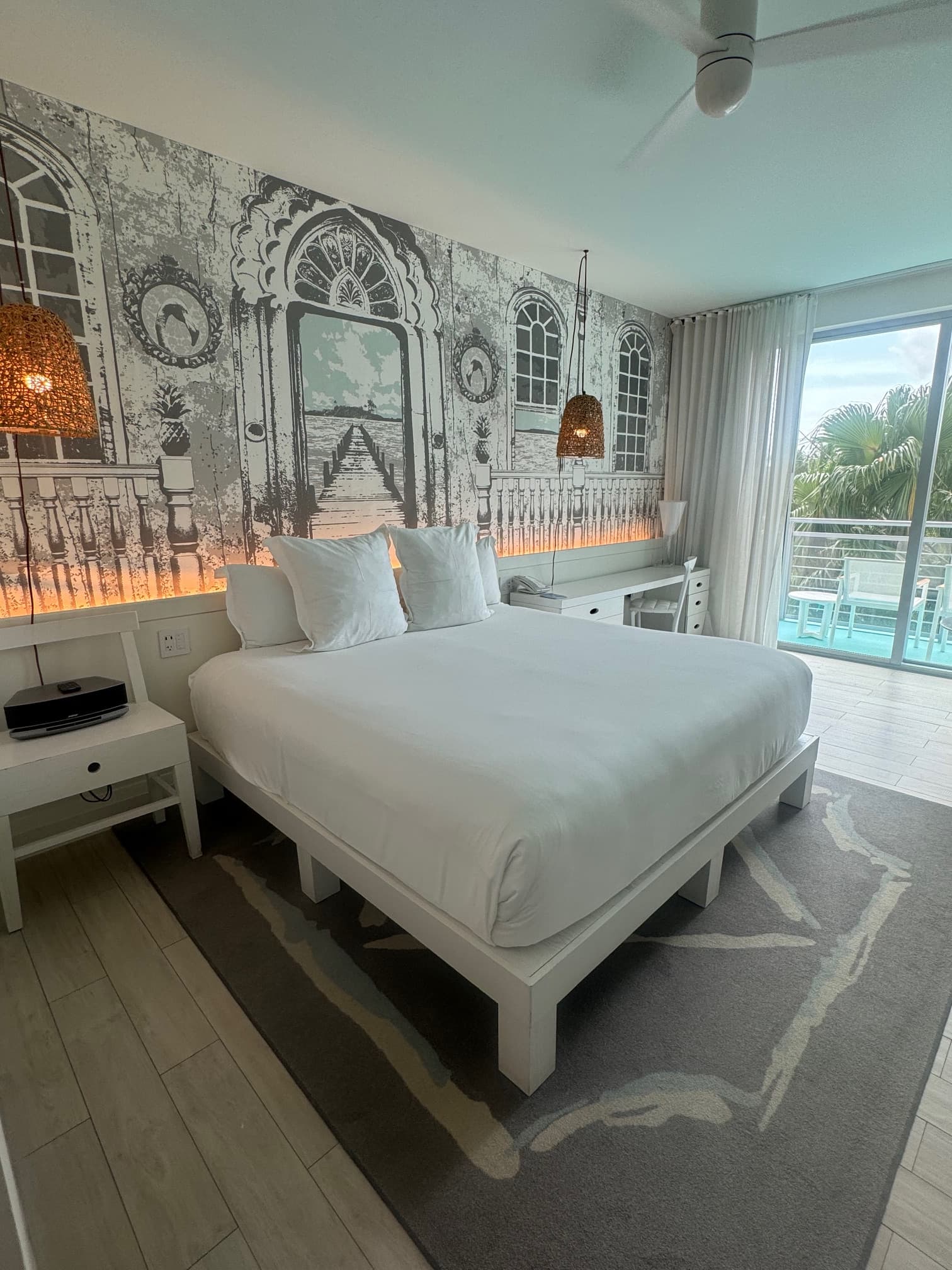 A hotel suite with a white bed, patterned rug, wallpaper, lamps, ceiling fan, side tables and view of the outside on the right side of the image.