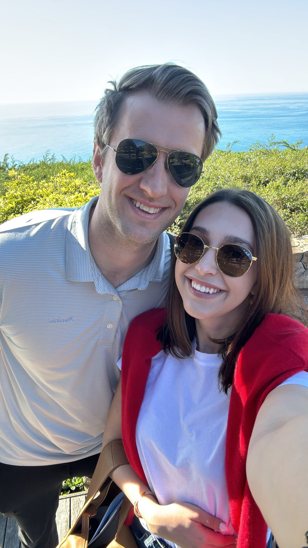 A couple posing in front of trees and the blue sea.
