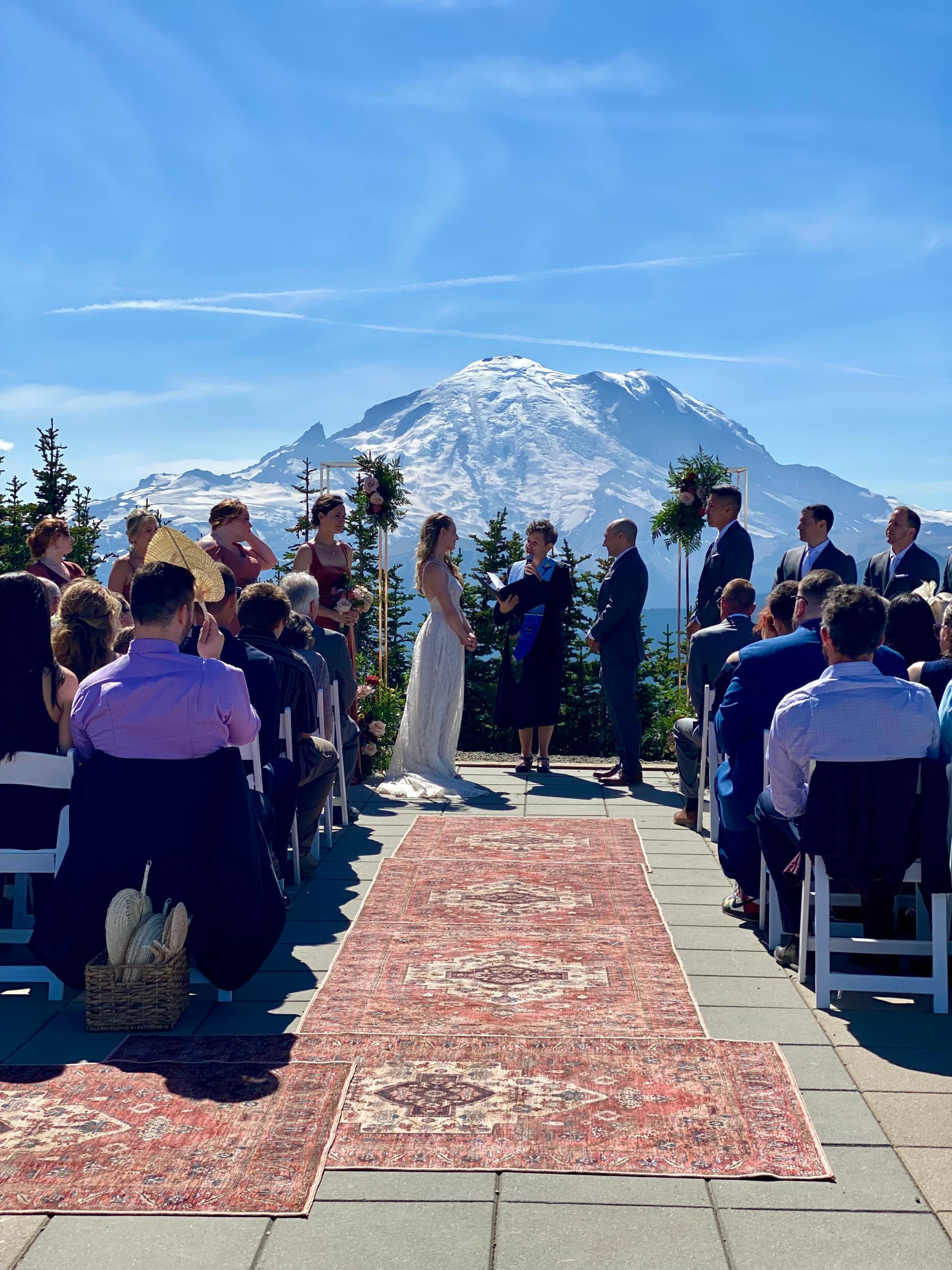 A couple getting married with guests seated on chairs, Turkish rugs lining a path to the altar and a couple joining hands with mountains in the background.