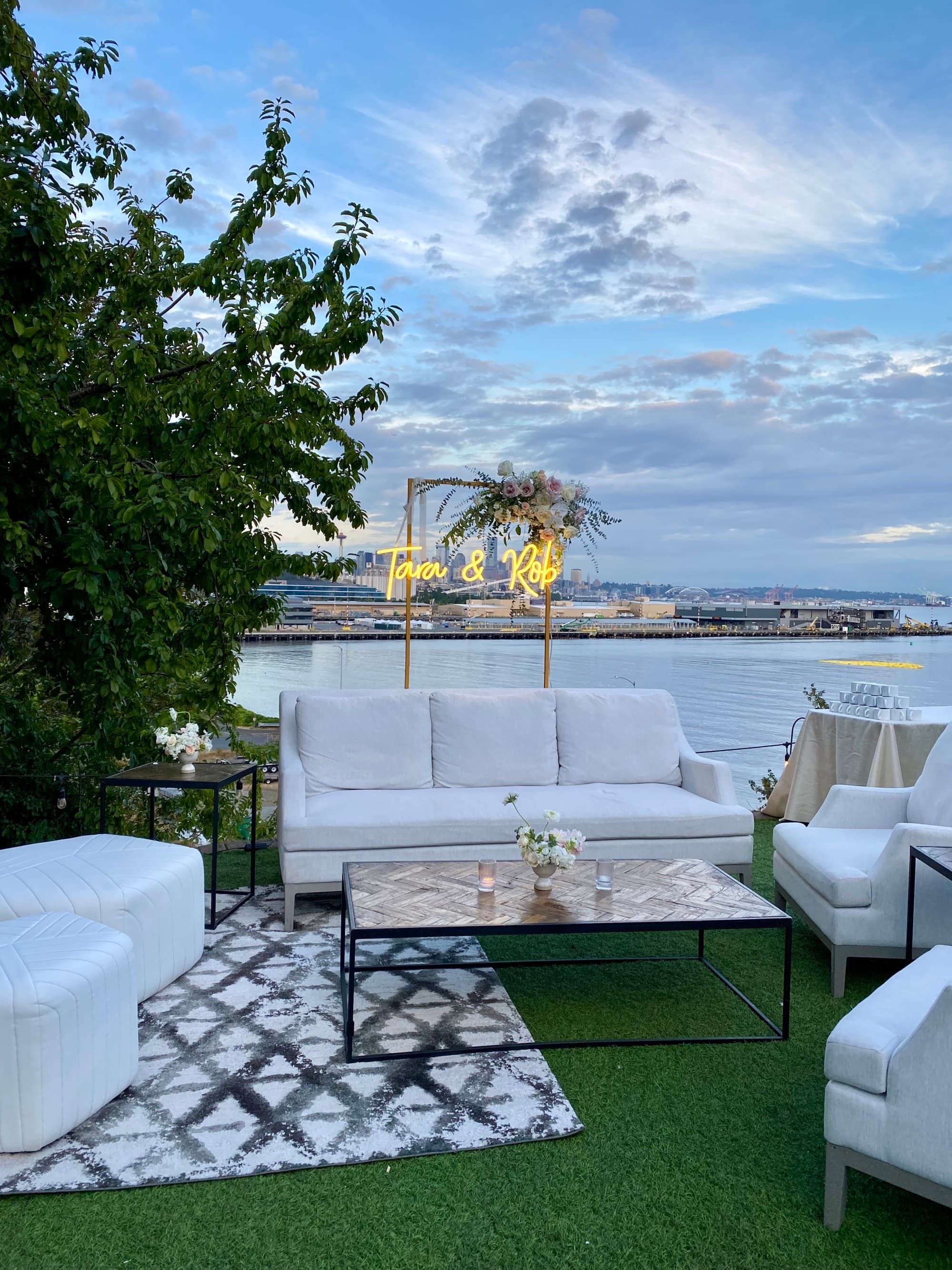 A terrace with couches, chairs and a coffee table outside. There are trees and a harbor in the background.