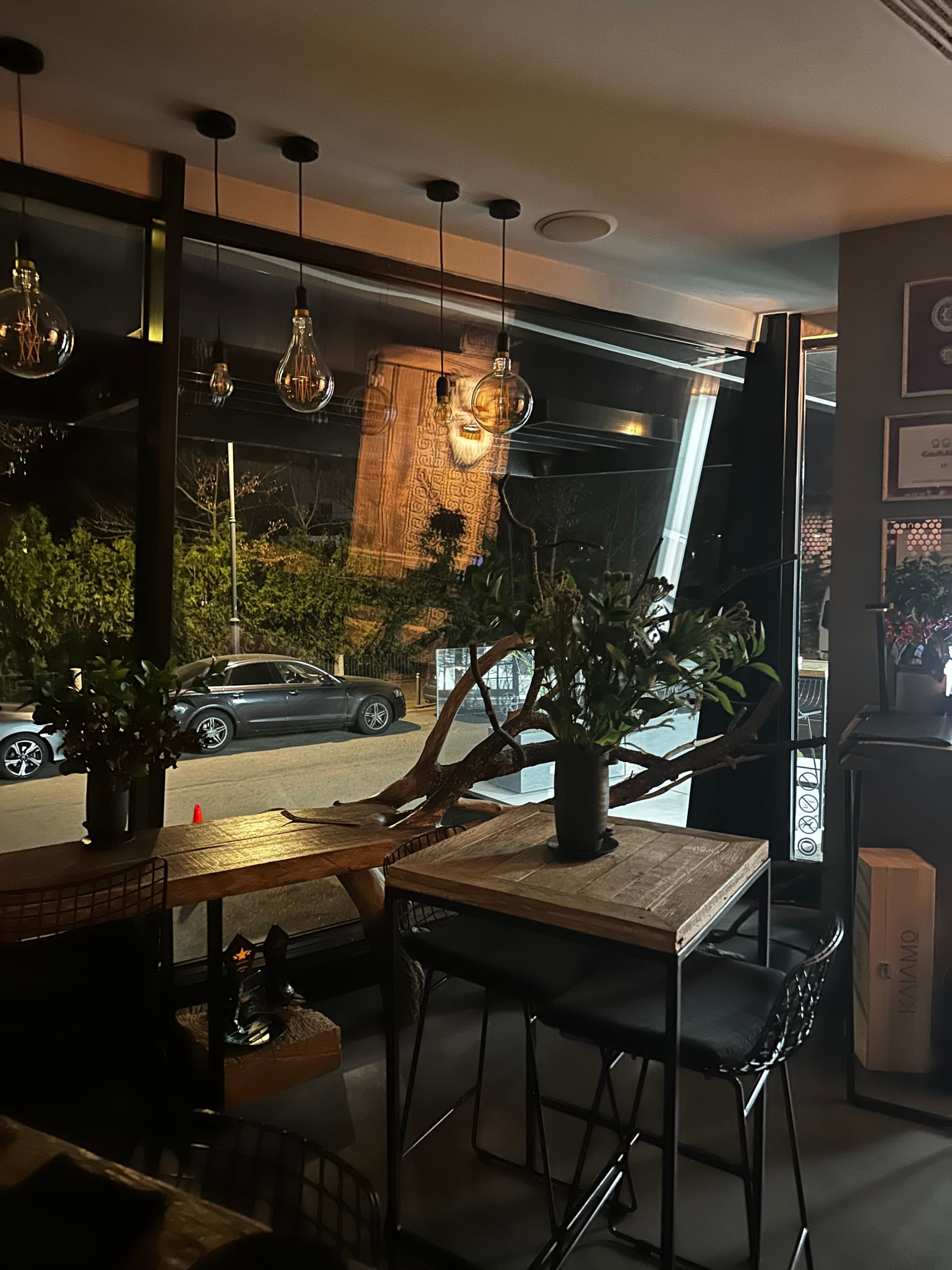 A picture of a restaurant interior with wooden high top tables, a window, hanging lights and trees.