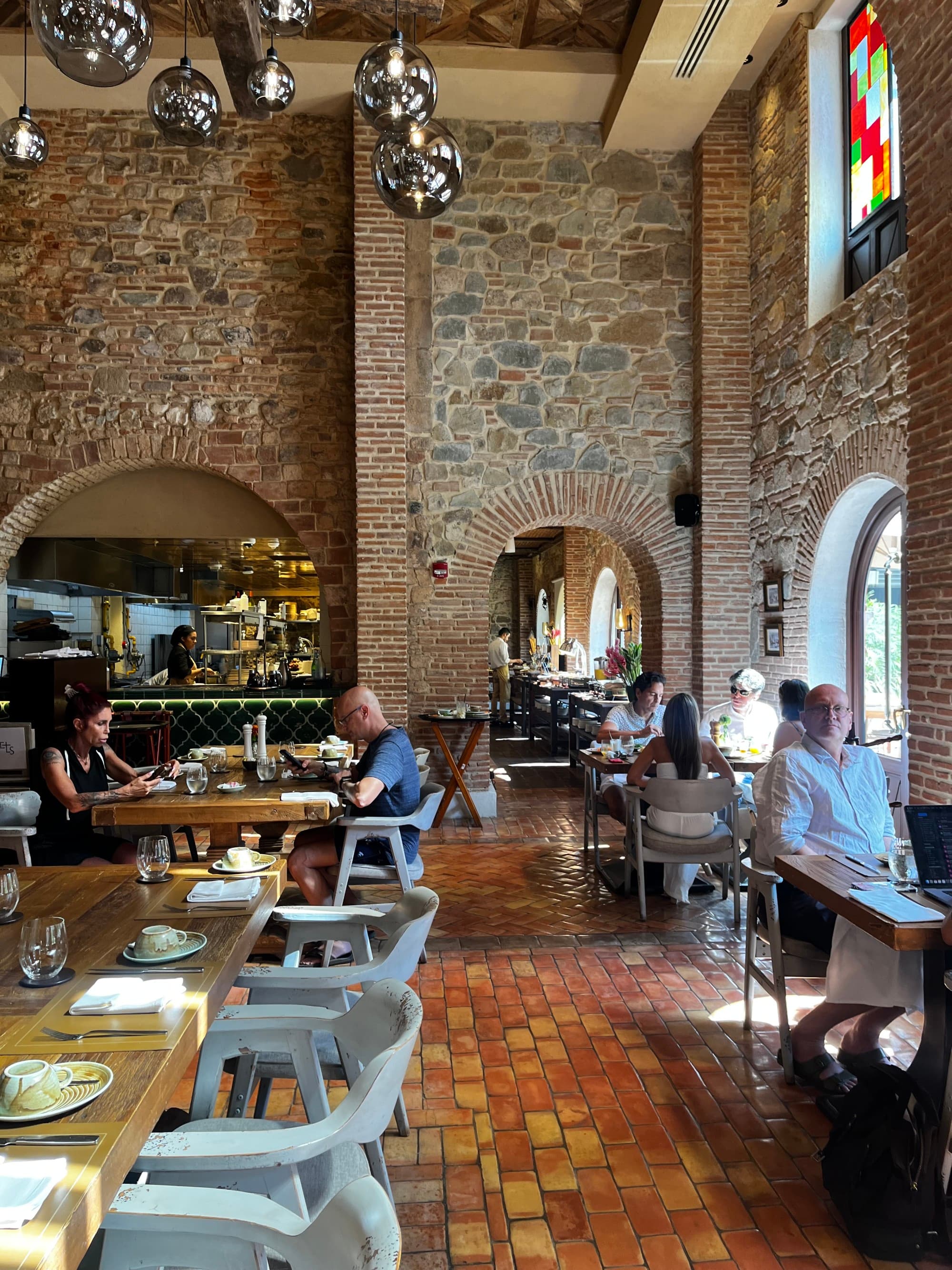 A restaurant interior with stone walls, a stained glass window, people dining at tables and silver pendant lighting.