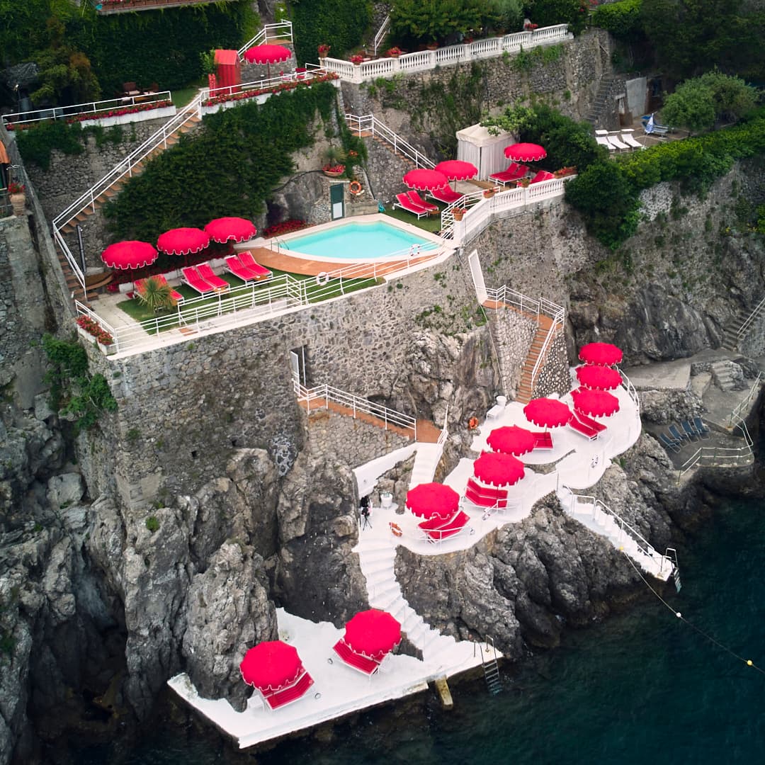 aerial view of a cliffside beachclub dotted with red umbrellas and red lounge chairs