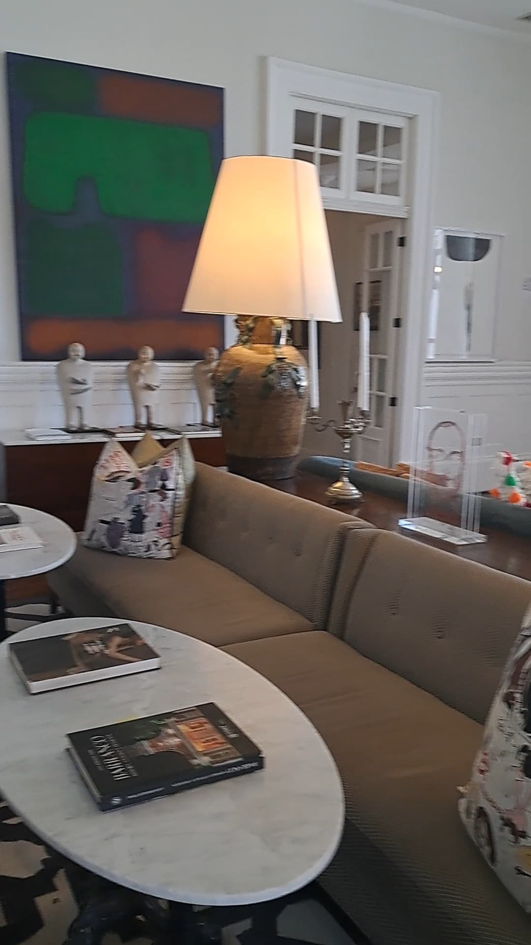 A hotel lobby complete with oval-shaped coffee tables, a beige couch, decorative pillows, a lamp, artwork and white walls.