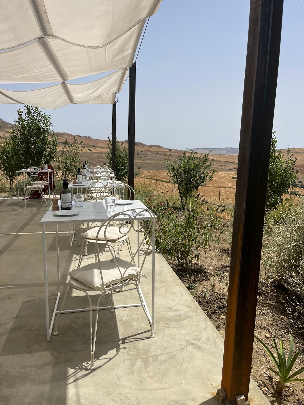 A patio with white tables and chairs, wooden beams and a white shade covering. There are bushes and desert-like terrain in the surrounding areas.