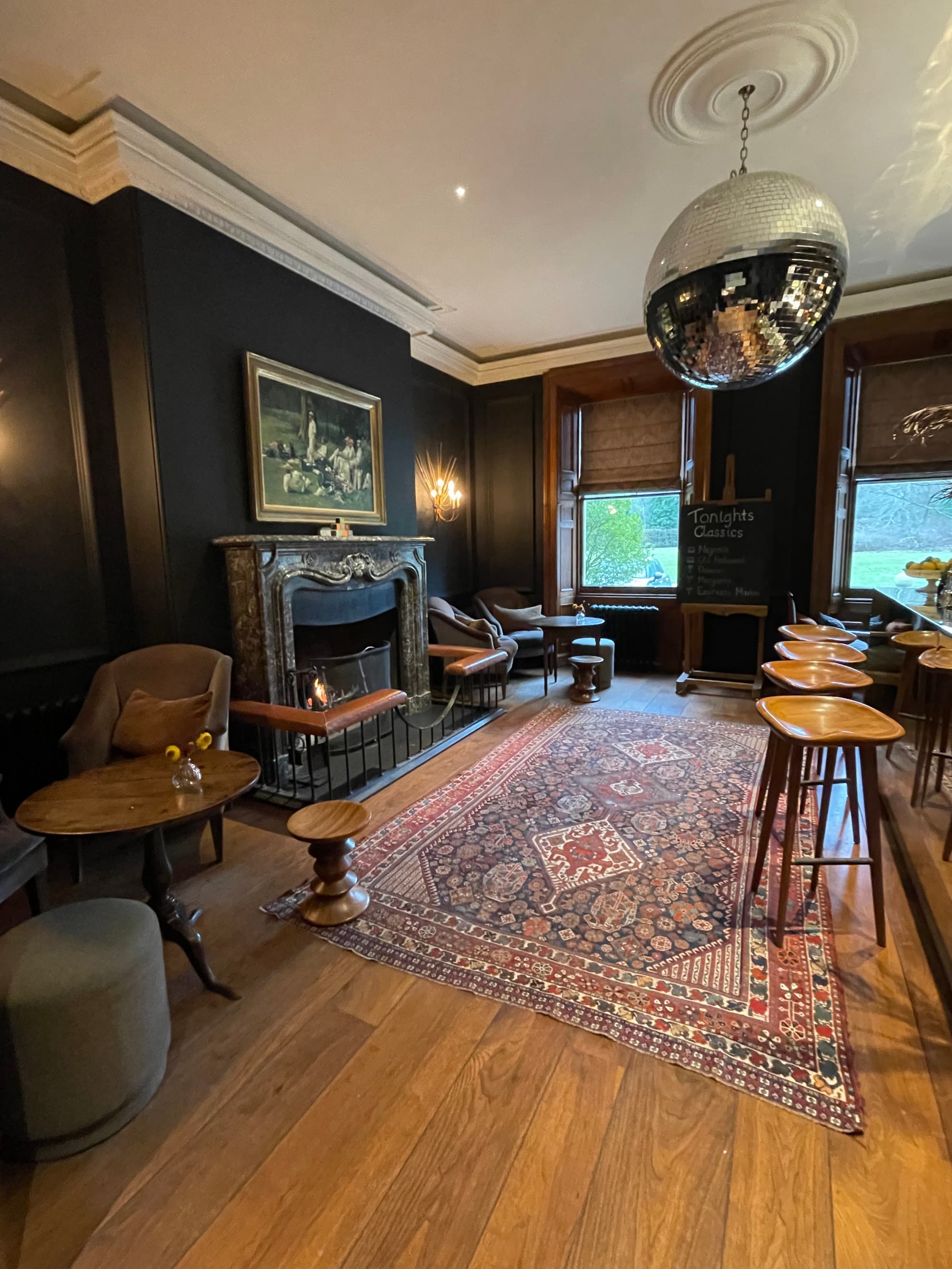 A cozy lounge with a fireplace, small table, rug, bar stools and windows looking onto a field