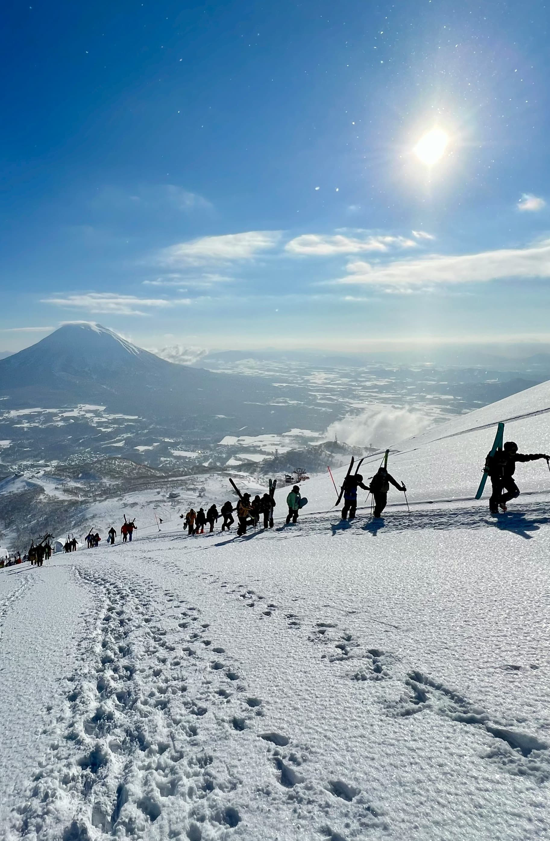 A group of people trekking up a snowy mountain with another large mountain in the far distance under the shining sun.