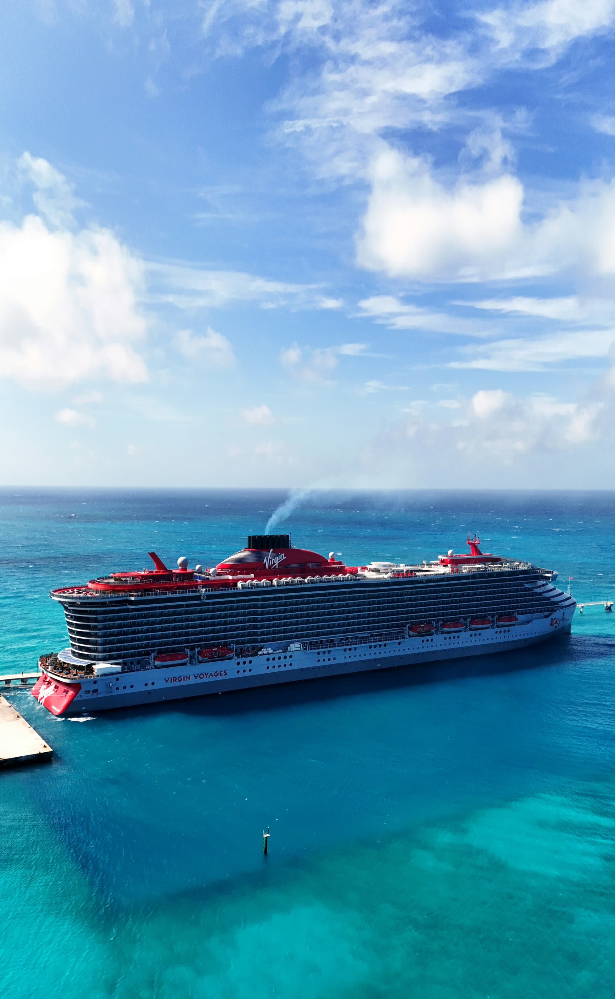 A Virgin Voyages cruise ship with a red top next to a dock in the ocean with puffy clouds in the blue sky