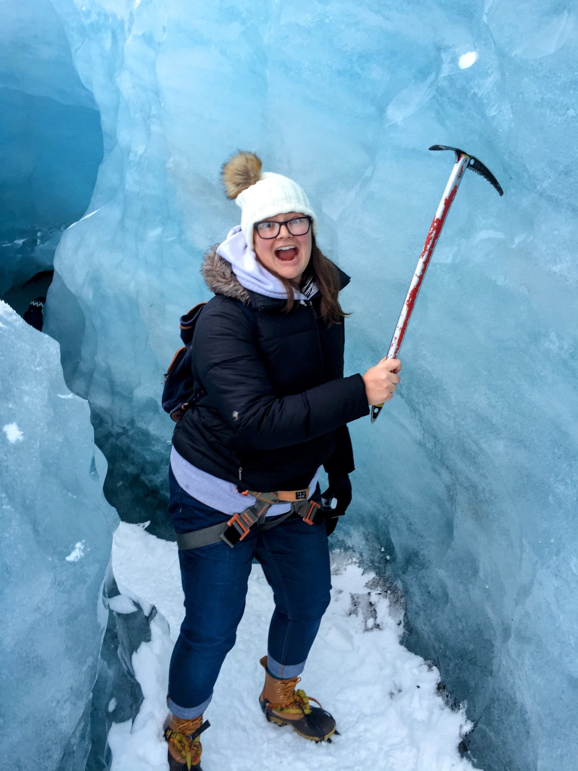 Posing with an ice pick near a large glacier wall and snow gear on.