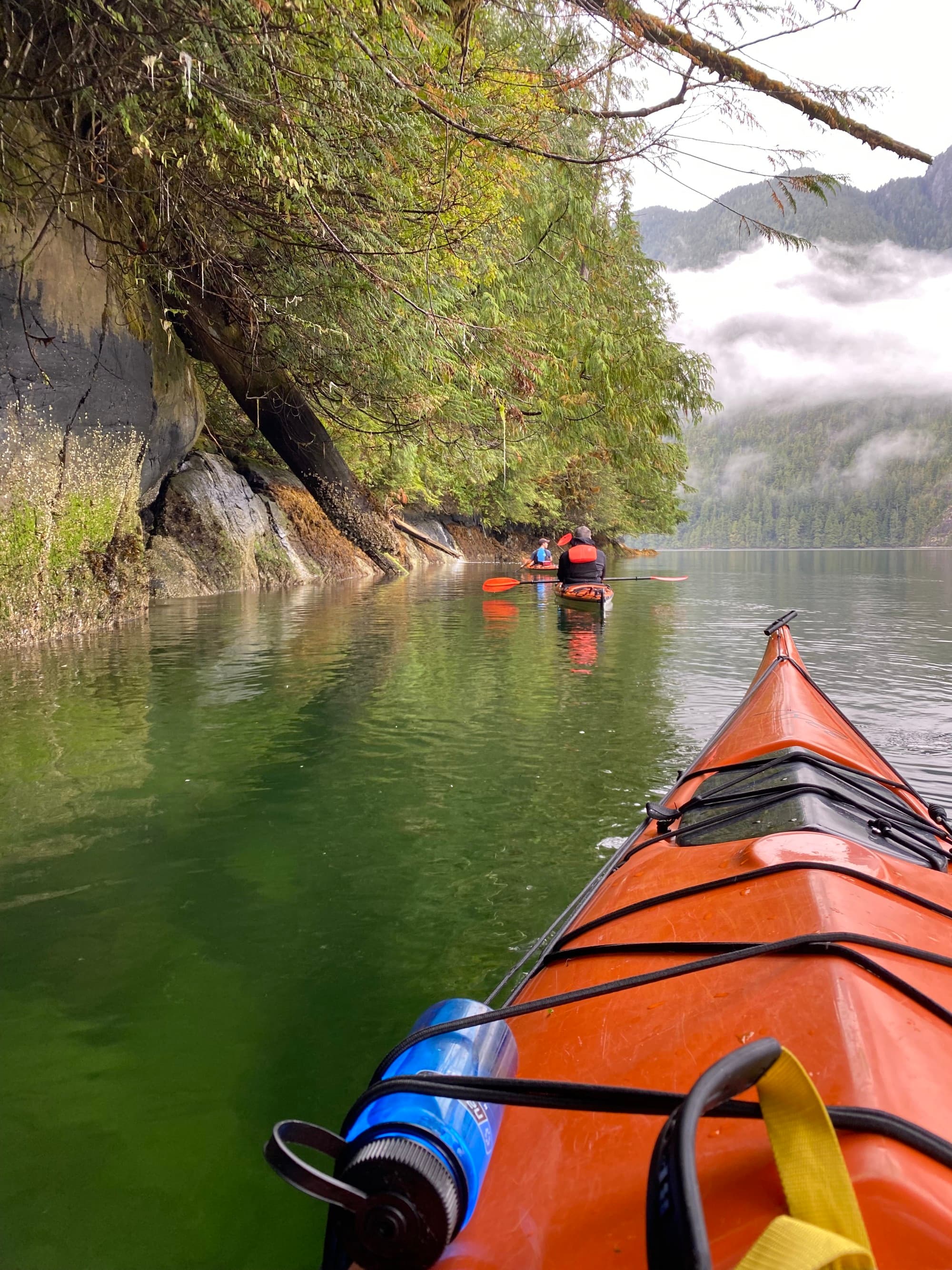 A view of an orange kayak in front of another orange kayak in the distance on top of green clear river water with trees and fog in the surrounding areas.