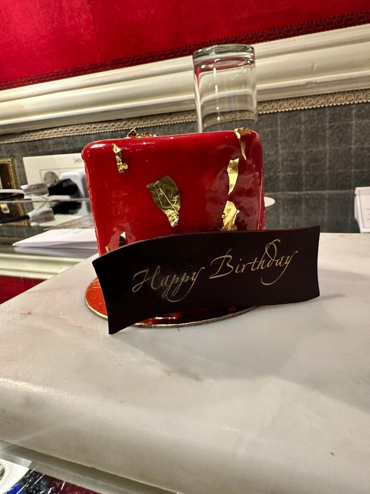 A red and gold foiled treat sitting on a marble countertop with a sign that reads 'Happy Birthday' in gold letters.