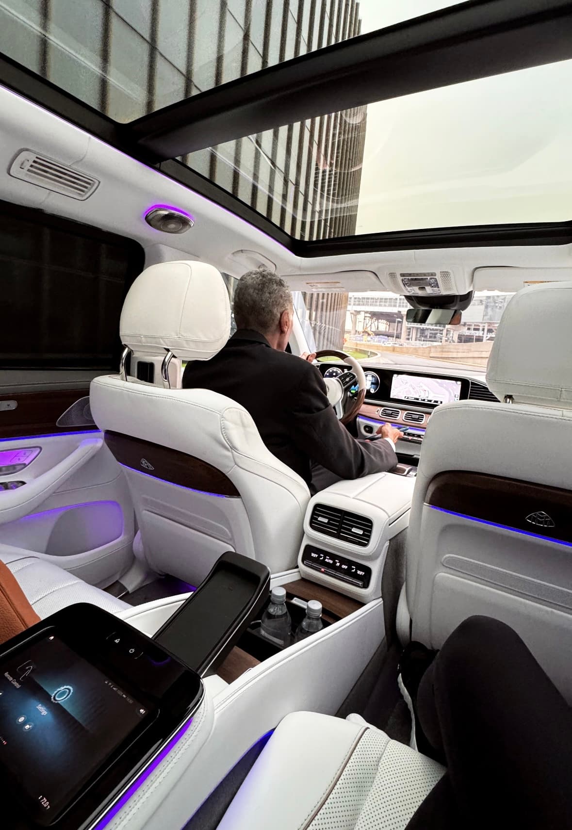 An interior view of a luxury car with purple lights, white leather seats and a driver in the front seat.