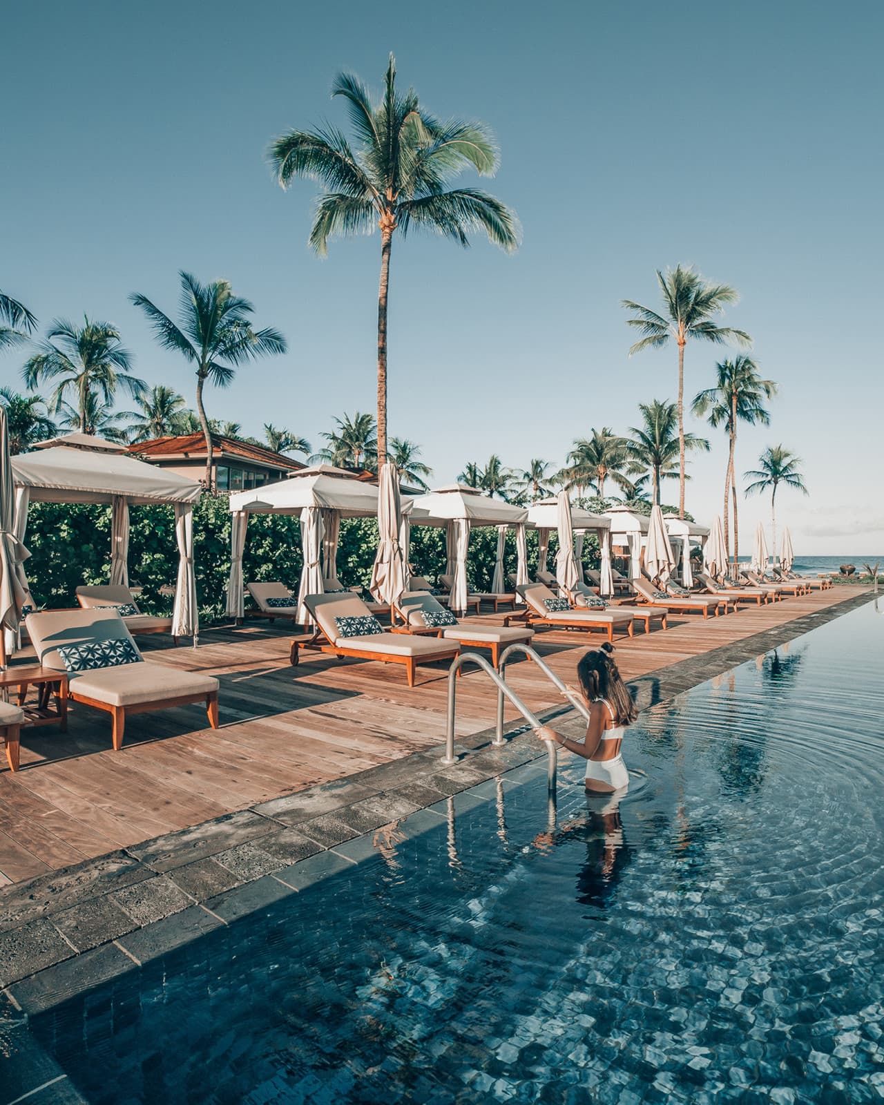 a woman in a white bathing suit steps out of a pool on a sunny day in the tropics