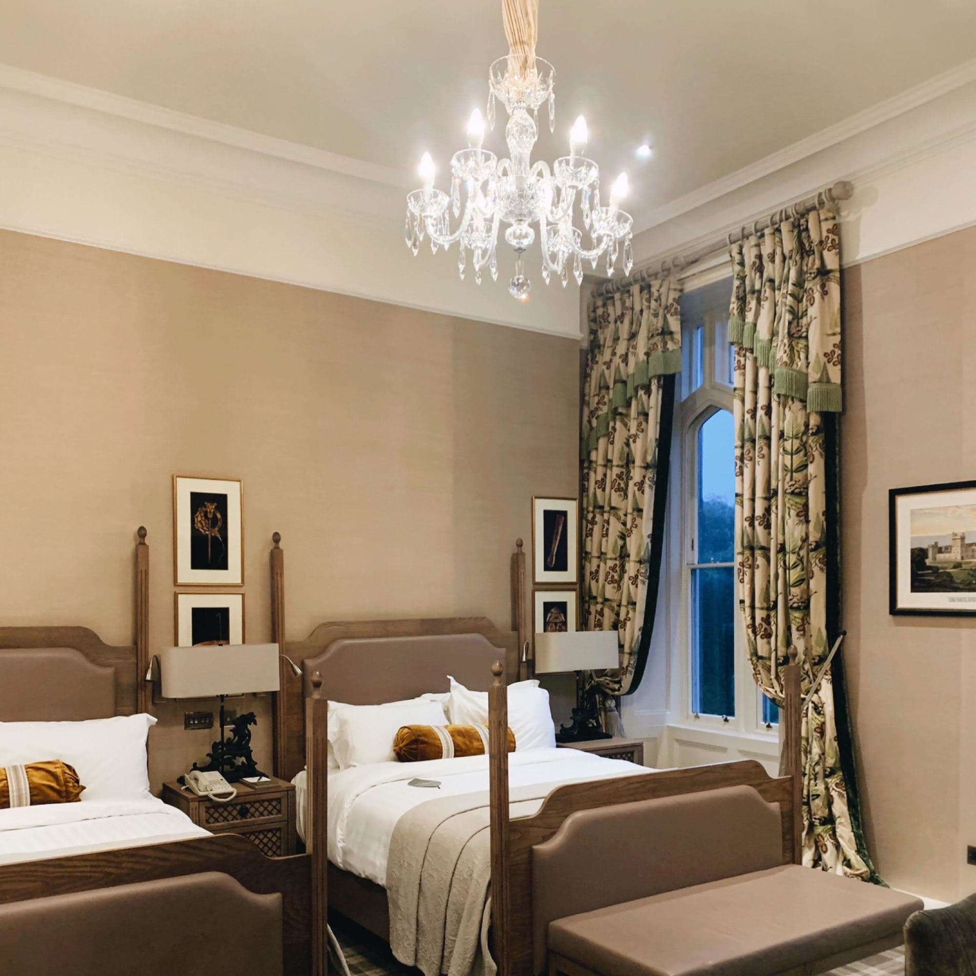 A two bed bedroom with a crystal chandelier, decorative curtains, artwork and pillows.